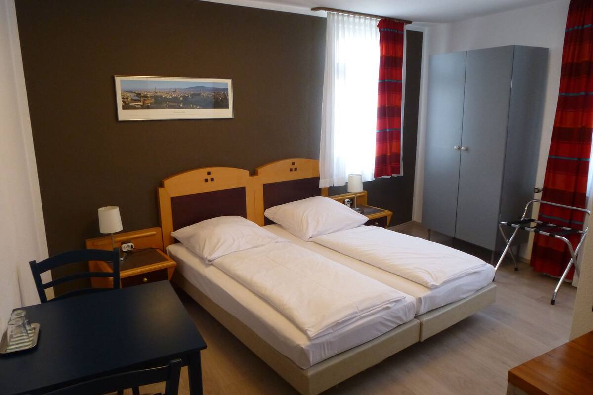 Habitación de hotel con dos camas individuales, una mesita de noche de madera con lámpara, una mesa con silla, un armario y una imagen en el marco con un paisaje urbano.