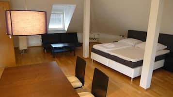 A compact room with a bed, a dining table with chairs, a sofa, and a lamp.
