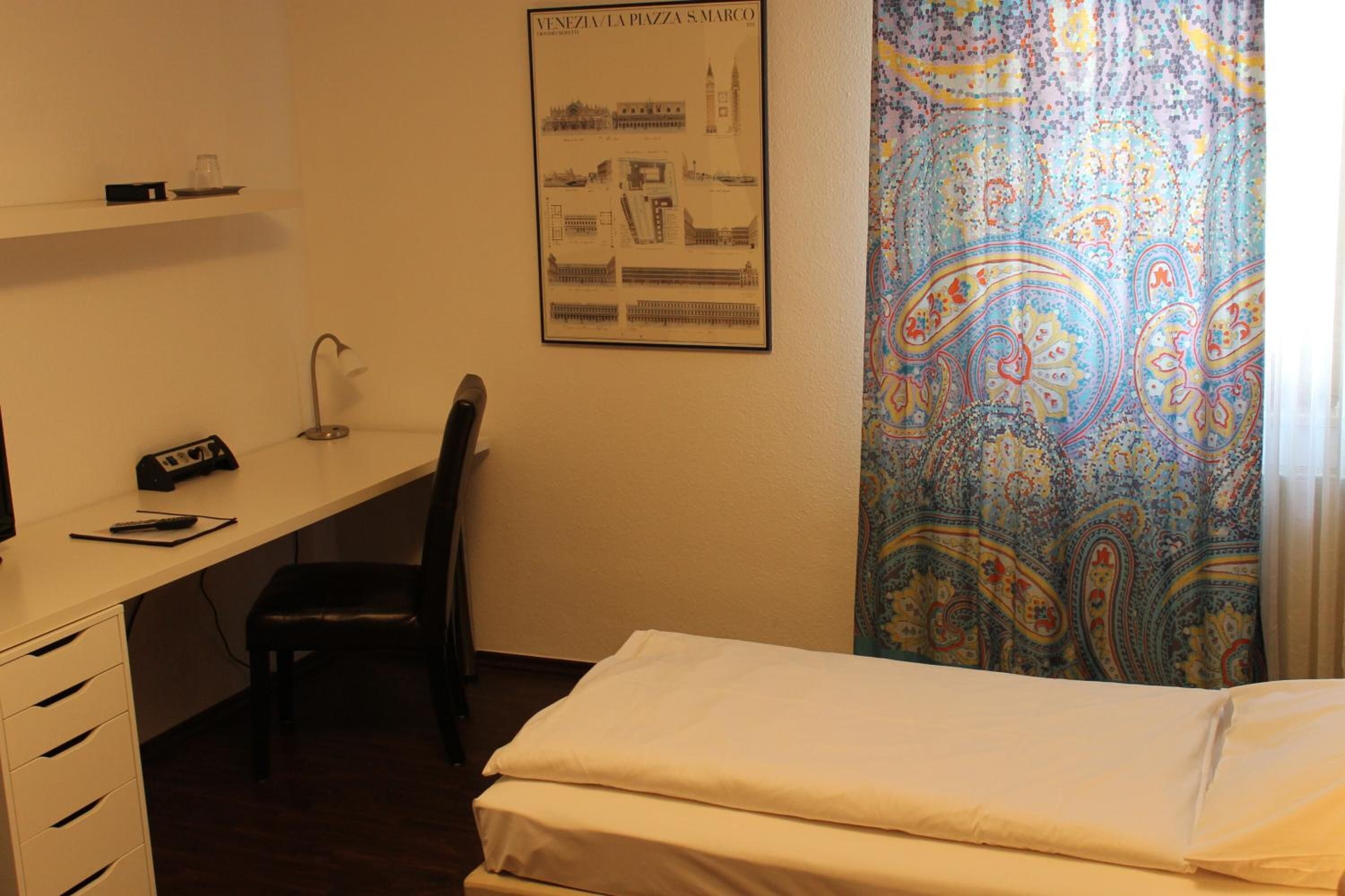 A hotel room with a bed, desk, chair, and a poster on the wall.