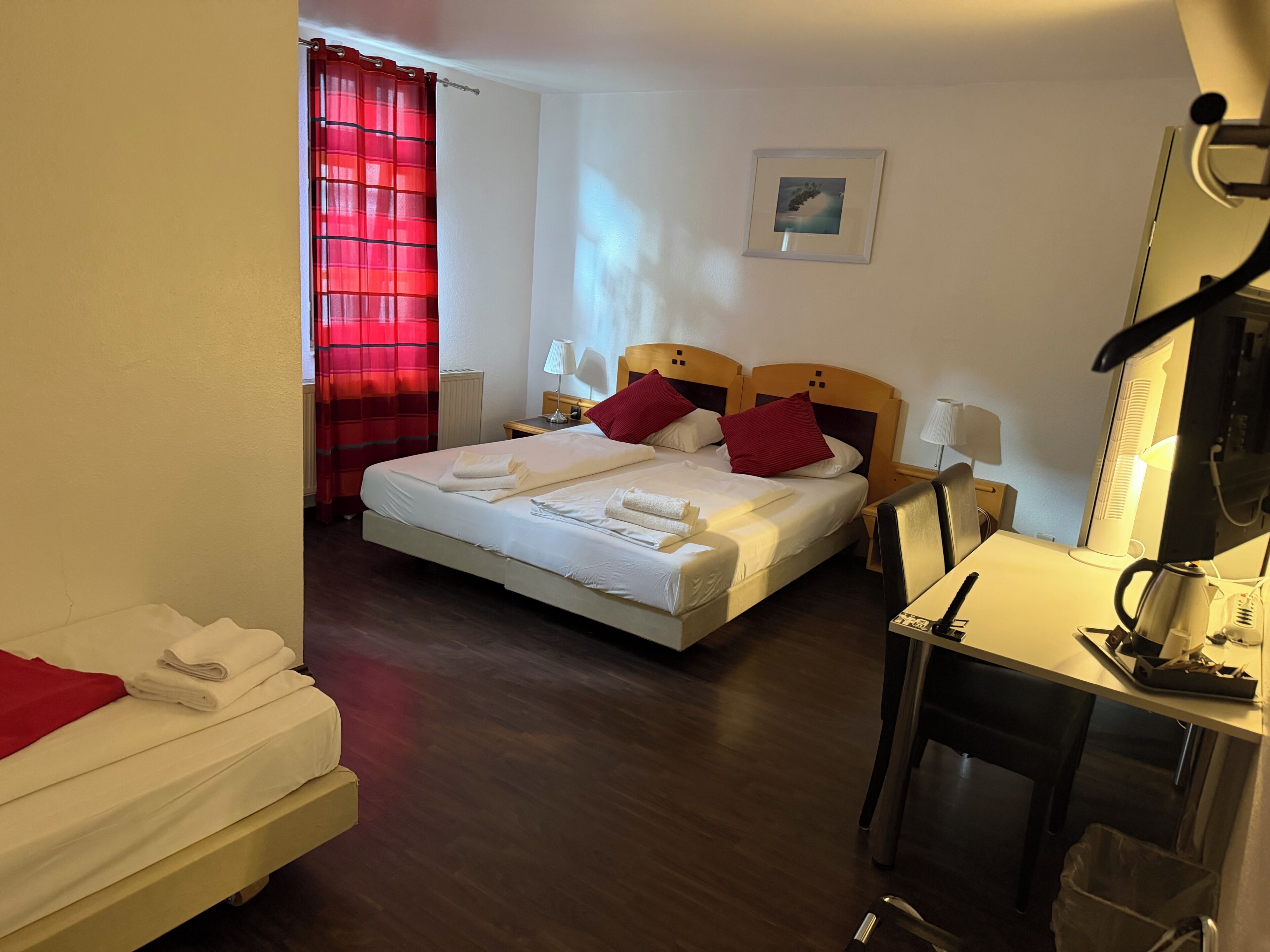 A hotel room with a bed, a desk, a chair, a lamp, a picture on the wall, and a red curtain.