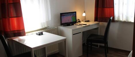A small hotel room with a white desk, a chair, a bed, a TV, and a lamp.