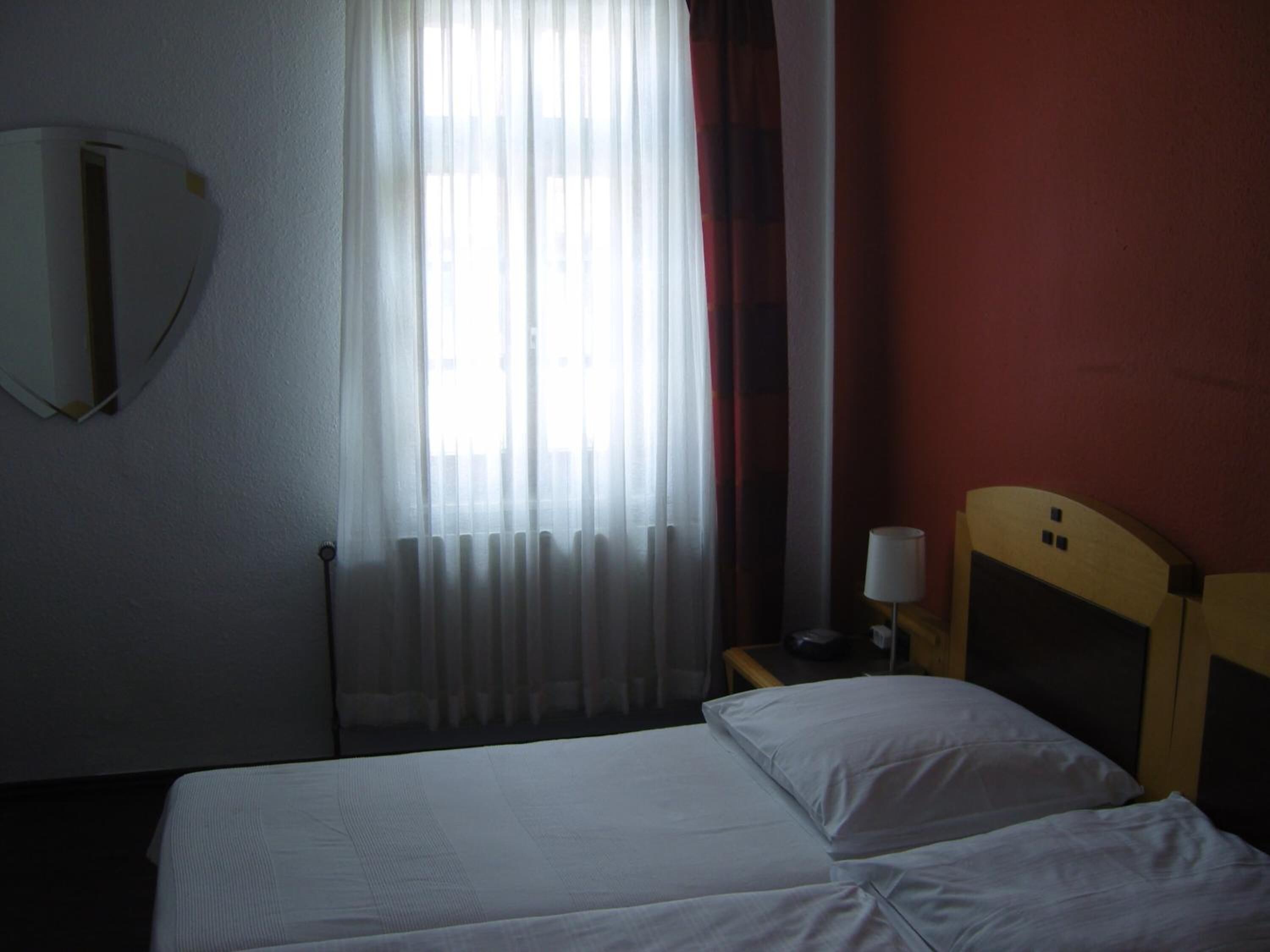 A hotel room with a bed, a bedside table with a lamp, a mirror, and a window with curtains.