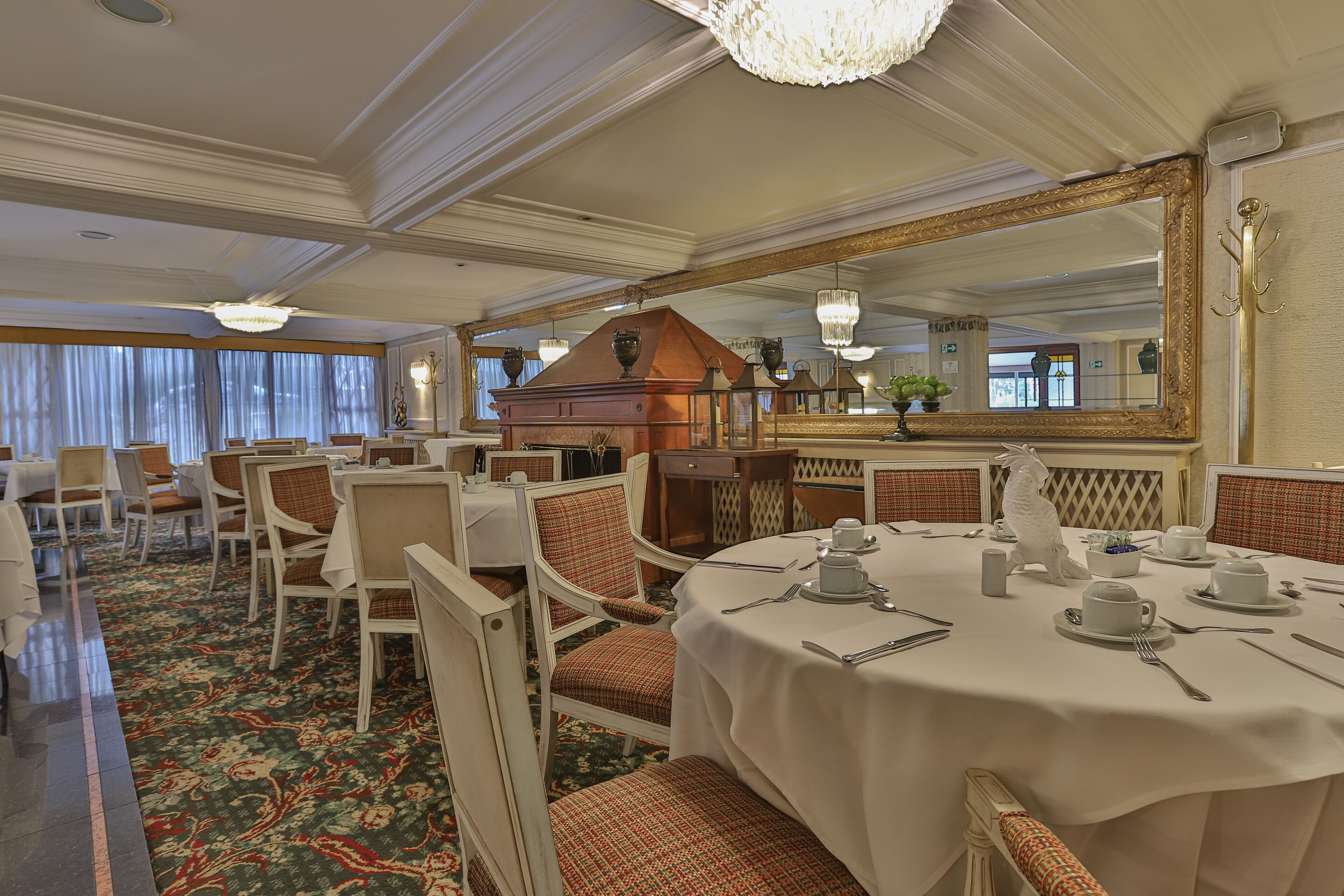 A dining area with round tables set for service, chairs with patterned cushions, a large ornate mirror, and a chandelier.