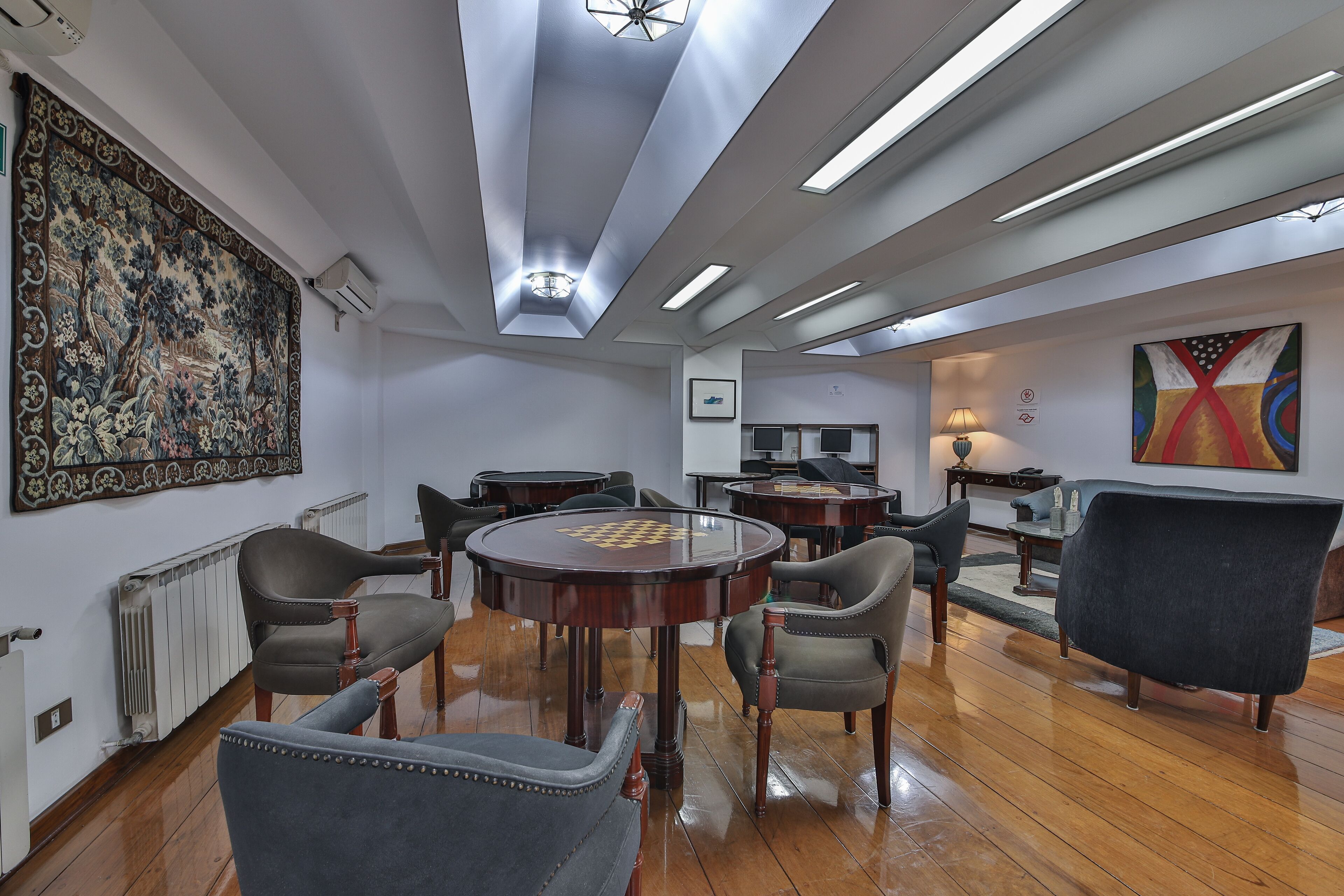 Una habitación amplia con un tapiz grande y ornamentado en la pared, una mesa redonda de madera con sillas, y un diseño de techo moderno con tragaluces.