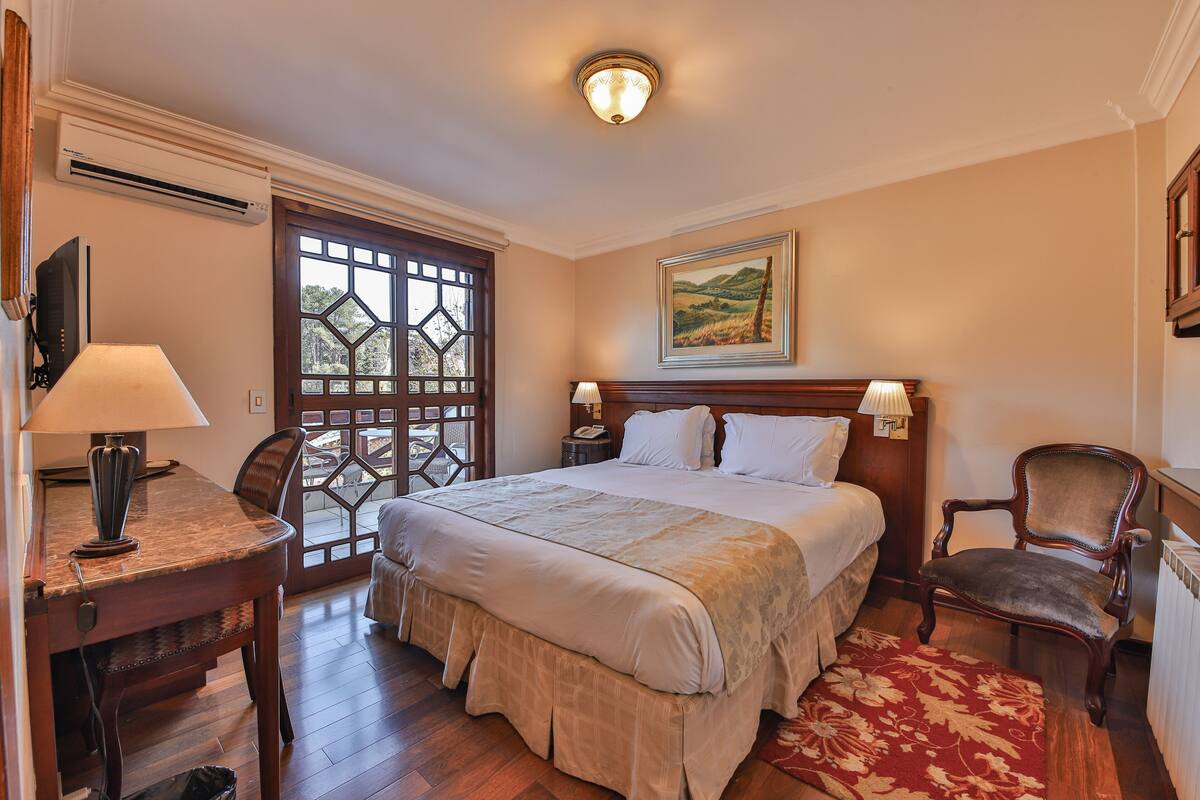 Ein Hotelzimmer mit einem großen Bett, einem Schreibtisch mit Lampe, einem Sessel, einem Bild an der Wand und einem Fenster mit Blick auf Grünflächen.