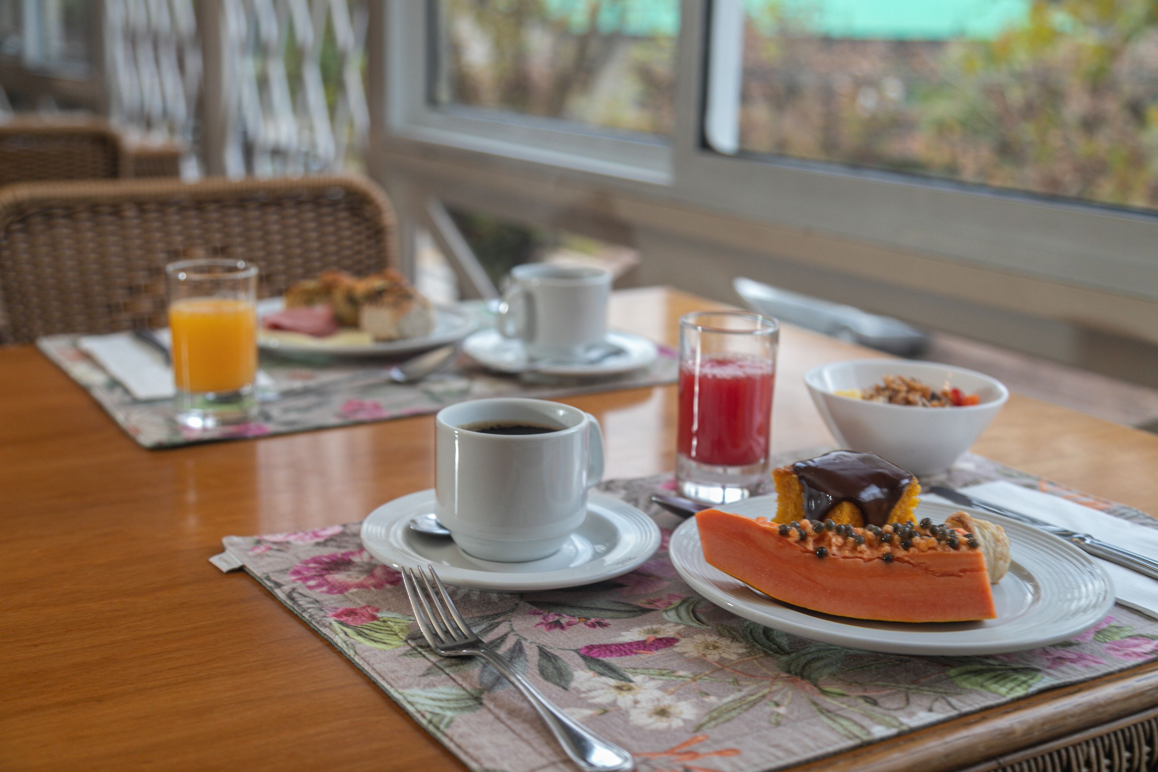Una mesa de desayuno con una taza de café, un vaso de jugo de naranja, un vaso de jugo rojo, un plato con un pastel y un tazón de cereal.