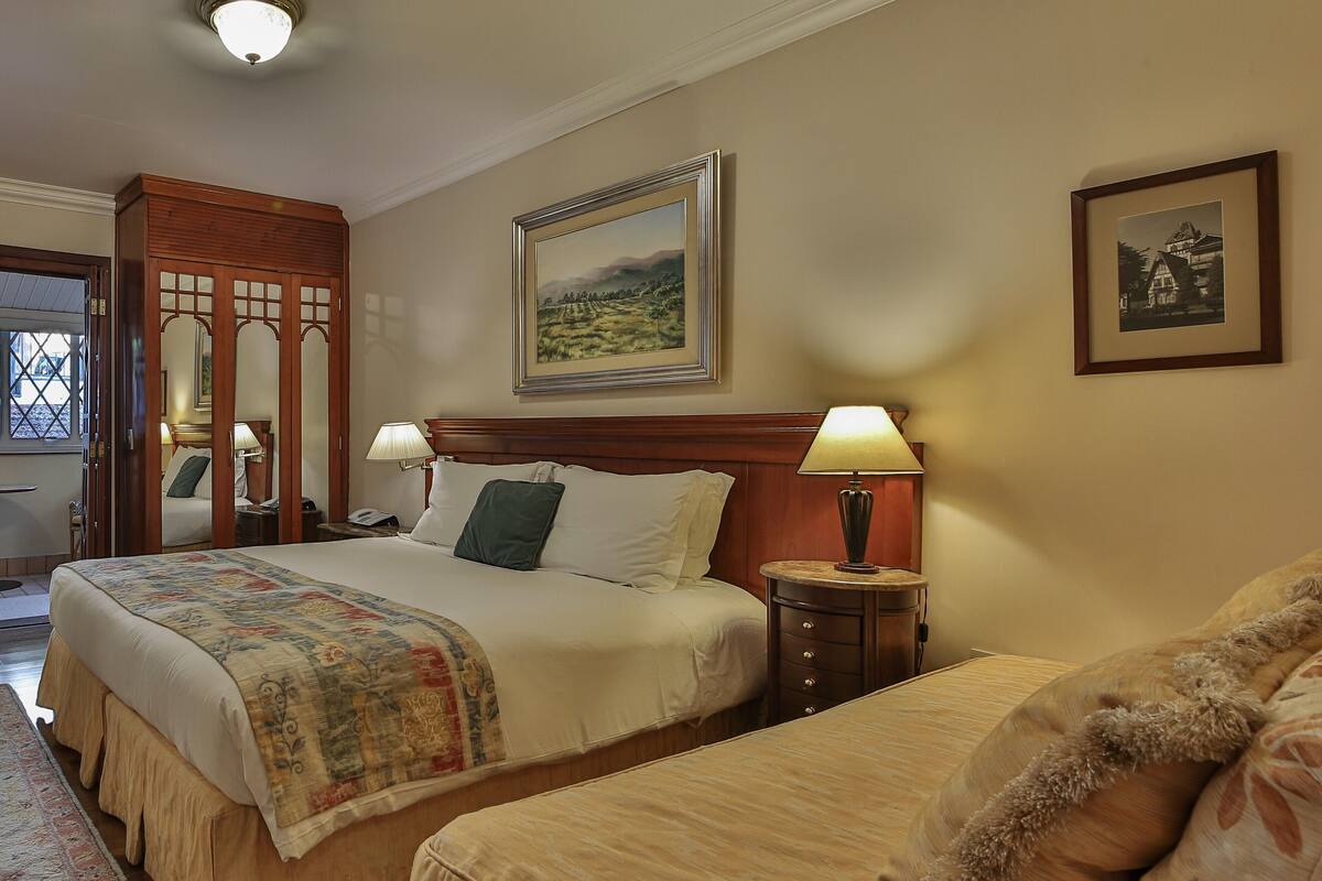 Ein Hotelzimmer mit einem großen Bett, einem kleineren Bett, einem Nachttisch mit Lampe, einem Holzschrank und einem Gemälde an der Wand.