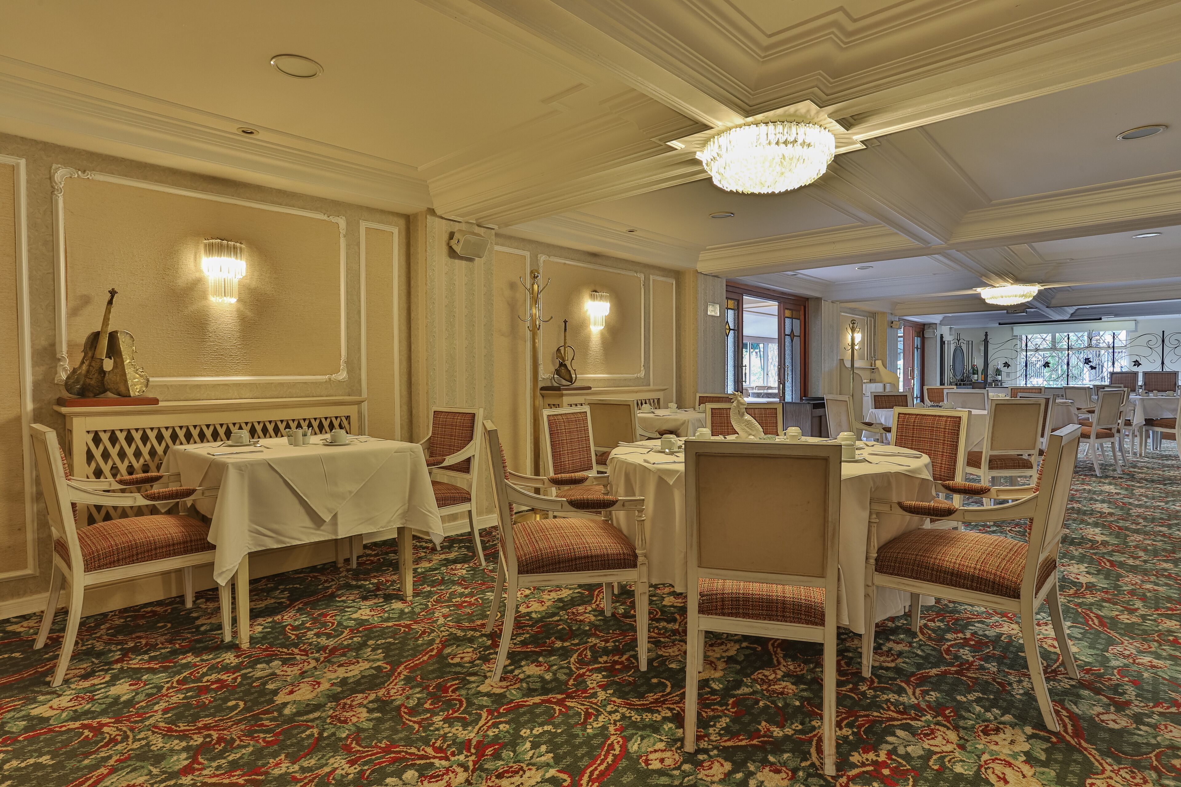 Uma área de jantar formal com mesas e cadeiras brancas, um tapete estampado e lustres.