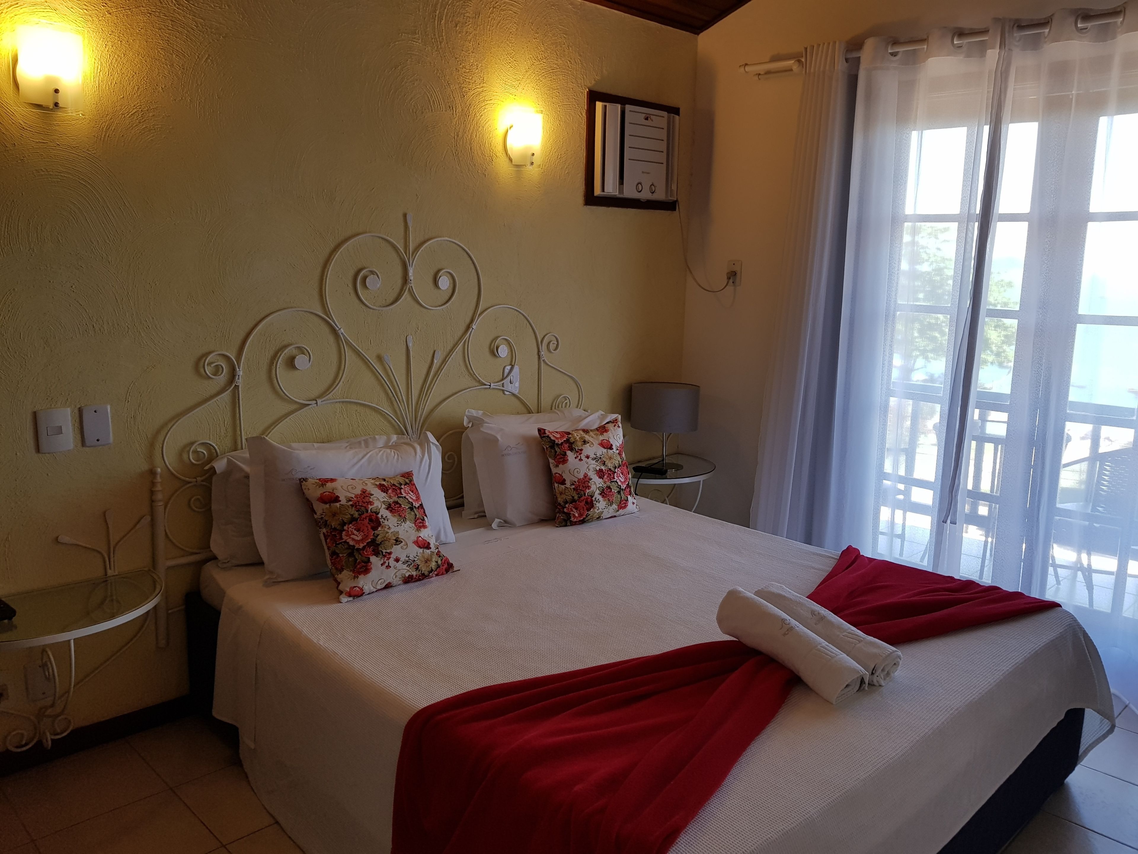 Una cama bien hecha con sábanas blancas y detalles rojos, una mesita de noche con lámpara y una ventana con cortinas traslúcidas.
