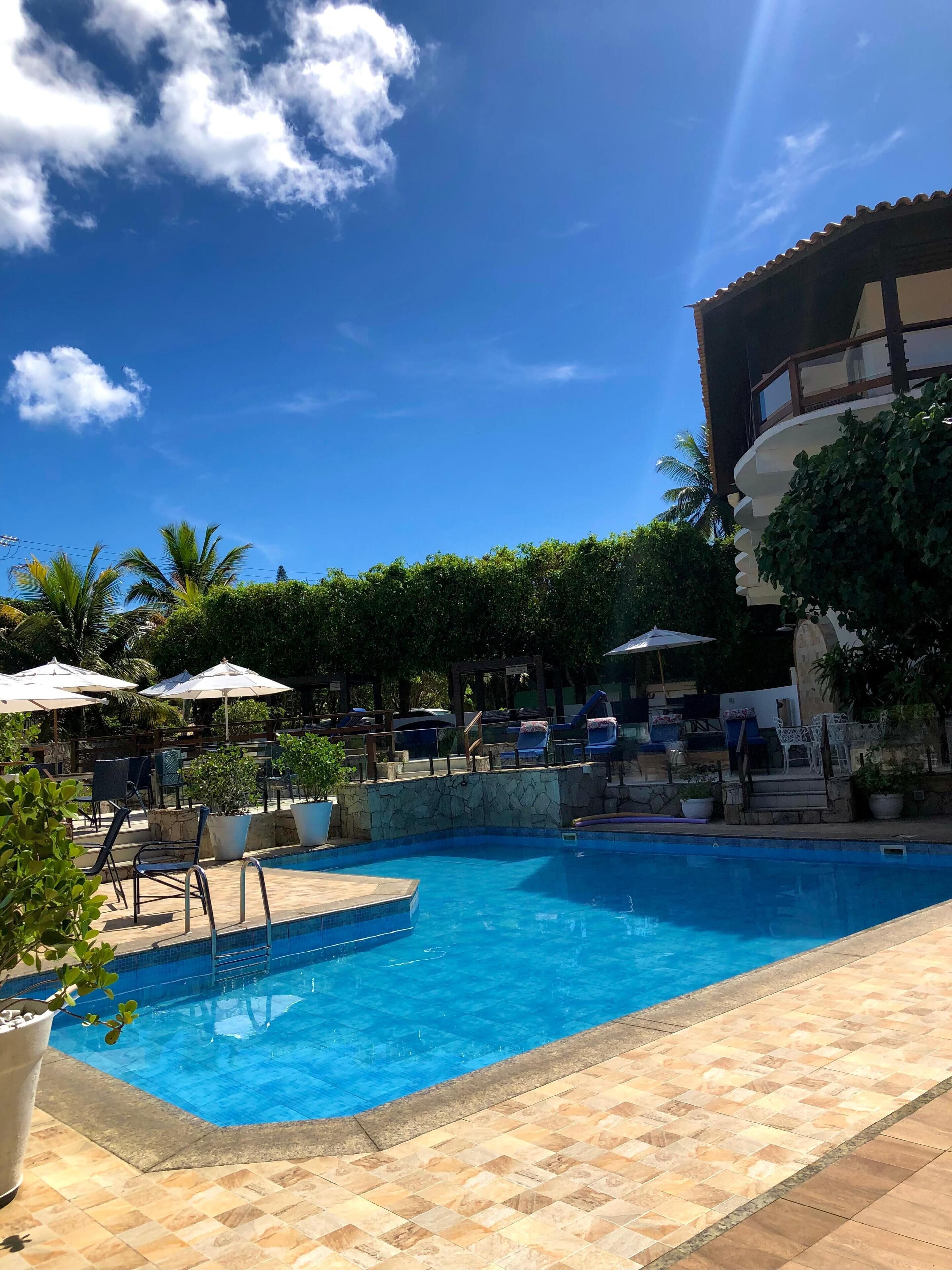 Una piscina con sillones de descanso y sombrillas, rodeada de vegetación y un edificio con balcón.