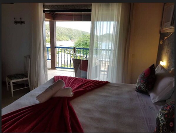 A hotel room with a bed, a chair, a balcony with a railing, and a view of greenery.