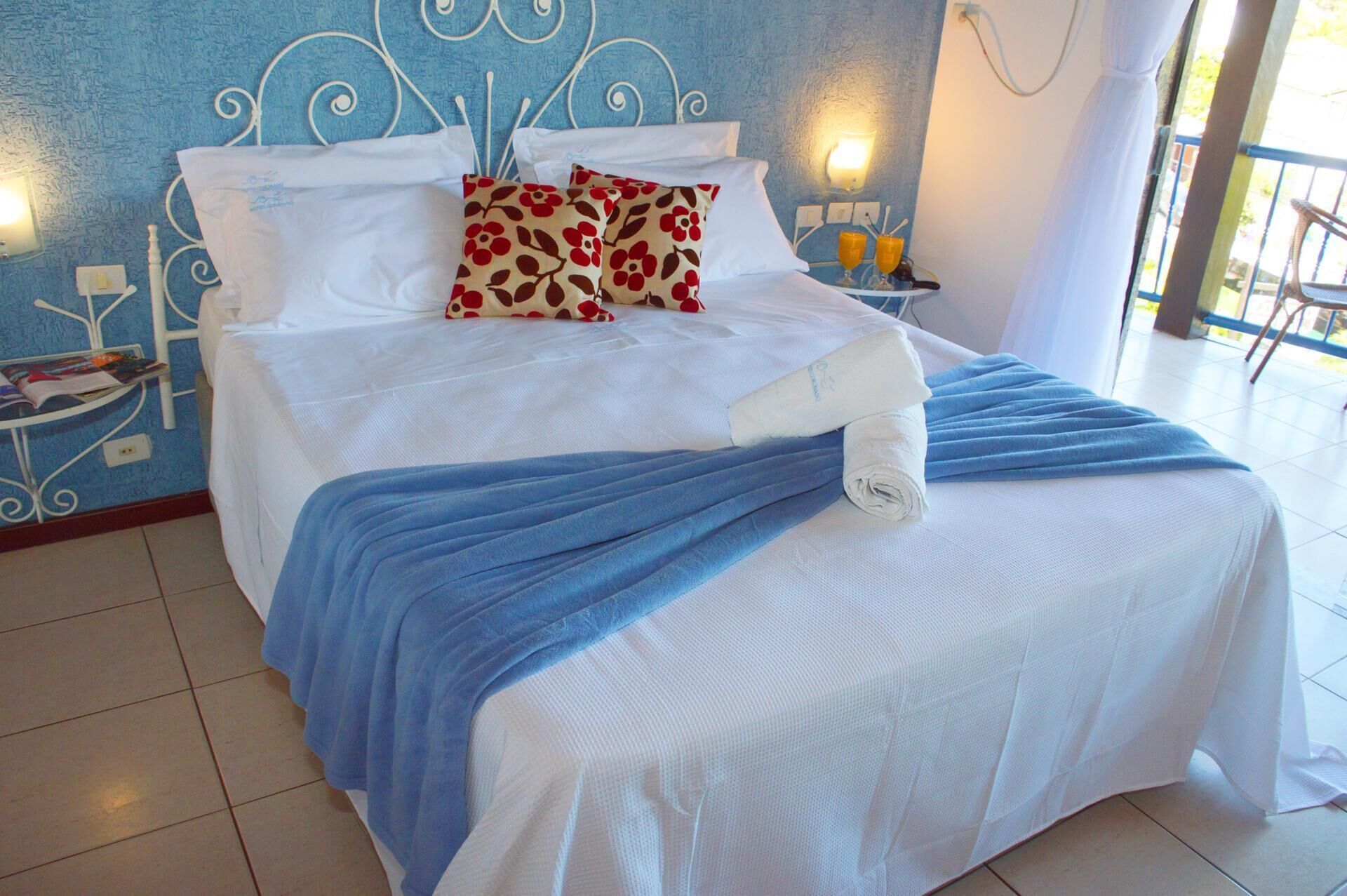A neatly made bed with white linens and a blue blanket, a floral pillow, and a white towel on the side.
