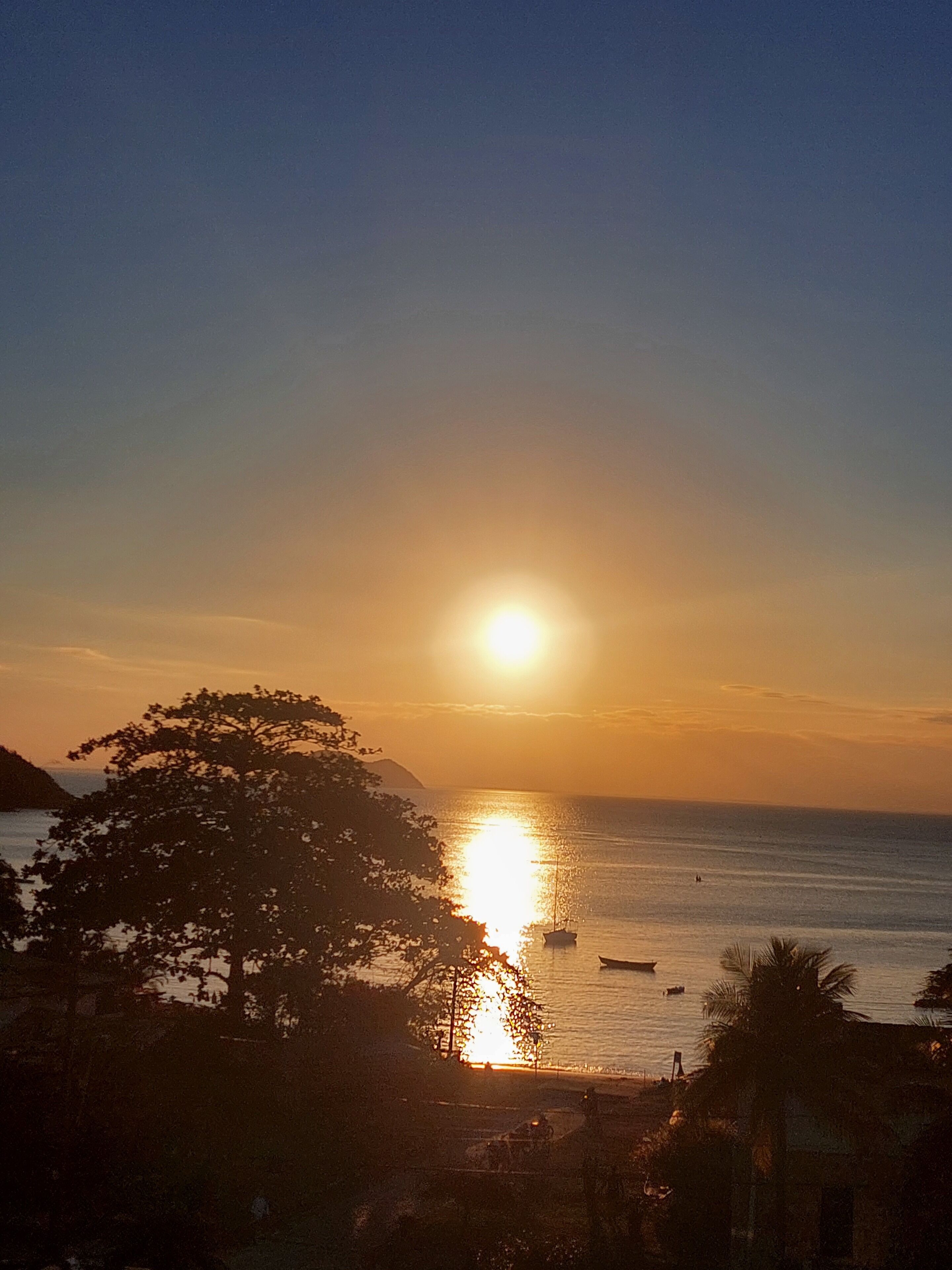 Un atardecer sobre un cuerpo de agua con barcos, árboles y una montaña distante.