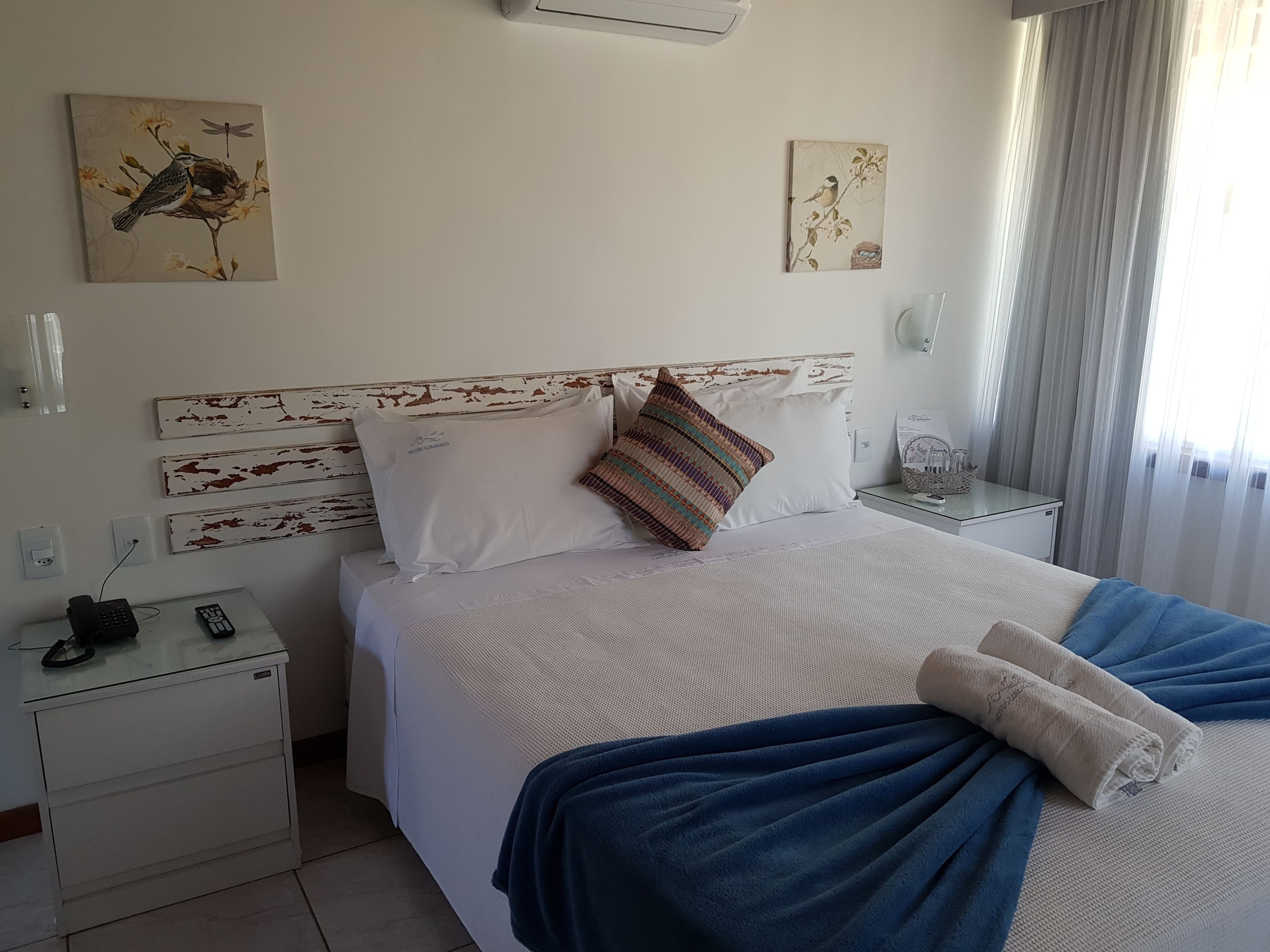 A neatly made bed with a blue blanket and a white towel, a bedside table with a phone and remote control, and wall art depicting birds and flowers.