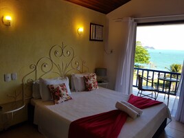 Un dormitorio con cama, almohadas, una manta roja, una mesita de noche, una lámpara y vistas al mar.