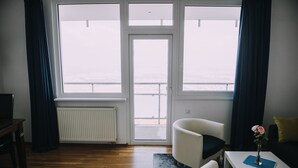 A room with a view of the sea through large windows, a white radiator, a wooden floor, a white chair, a small table with a vase of flowers, and a yellow pillow on a black sofa. - Hotel Bellevue (Kranj)