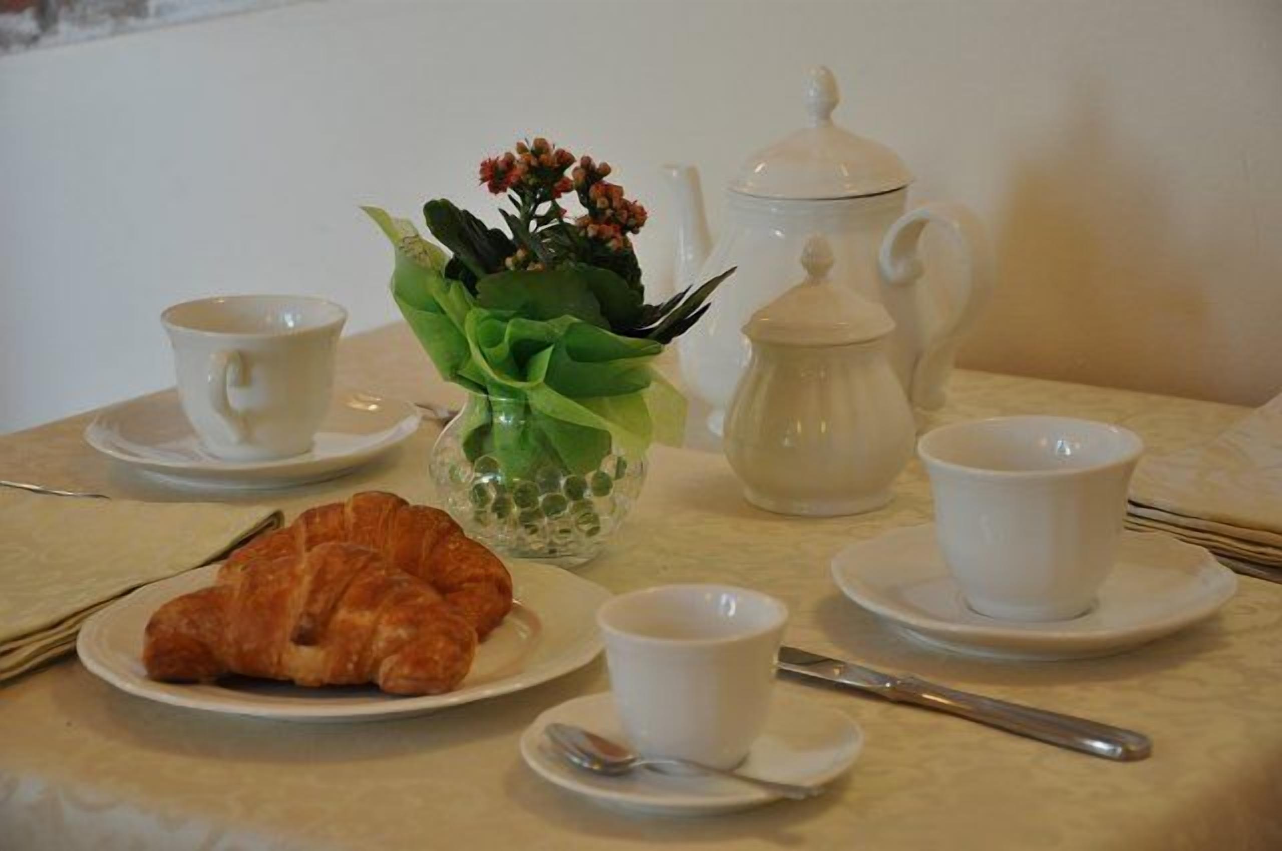 A table set with a croissant, two cups, a teapot, and a vase with flowers.