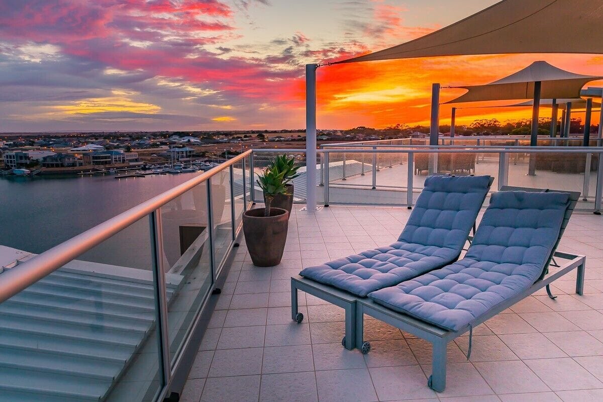 A balcony with two cushioned chairs, a potted plant, and a view of a marina during sunset.