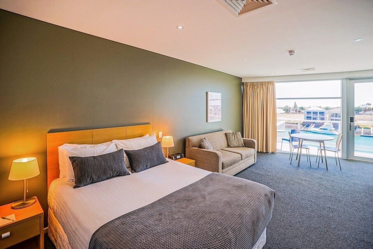 A hotel room with a large bed, a sofa, a dining table, and a view of a body of water through large windows.