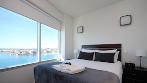Hotelkamer met een groot bed, nachtkastje, lamp en uitzicht op het water en de skyline.