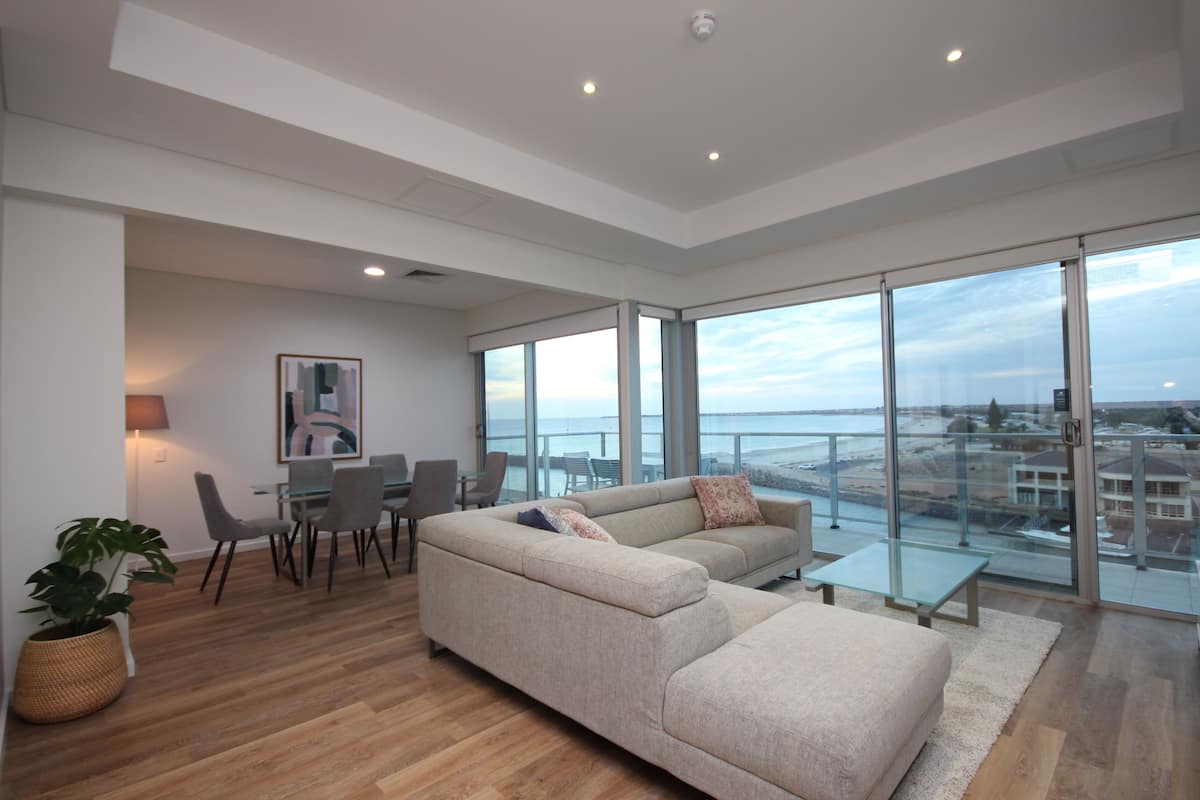 A modern living room with a large sofa, a dining area, and a view of the sea.