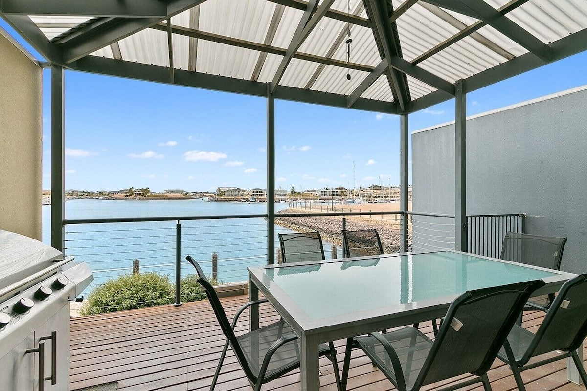 A covered outdoor patio with a glass table and chairs, overlooking a body of water and a marina.