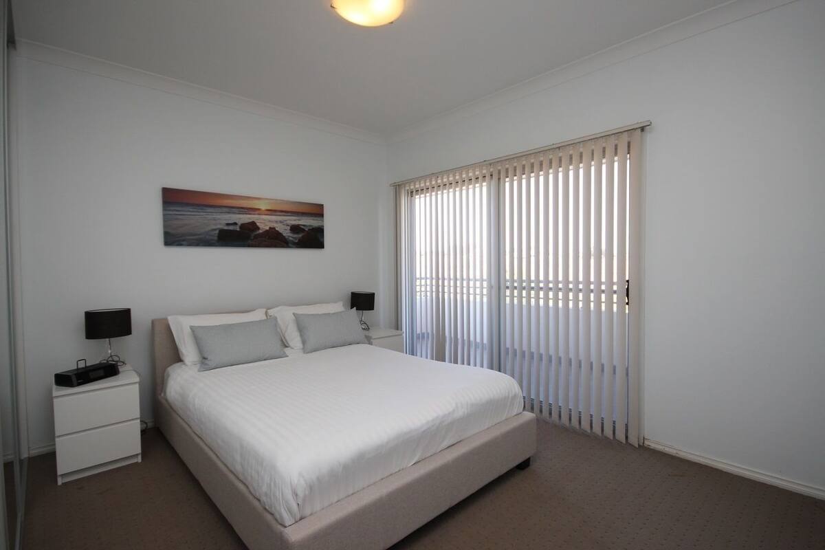 A bedroom with a bed, a nightstand, a lamp, a painting, and a window with blinds.