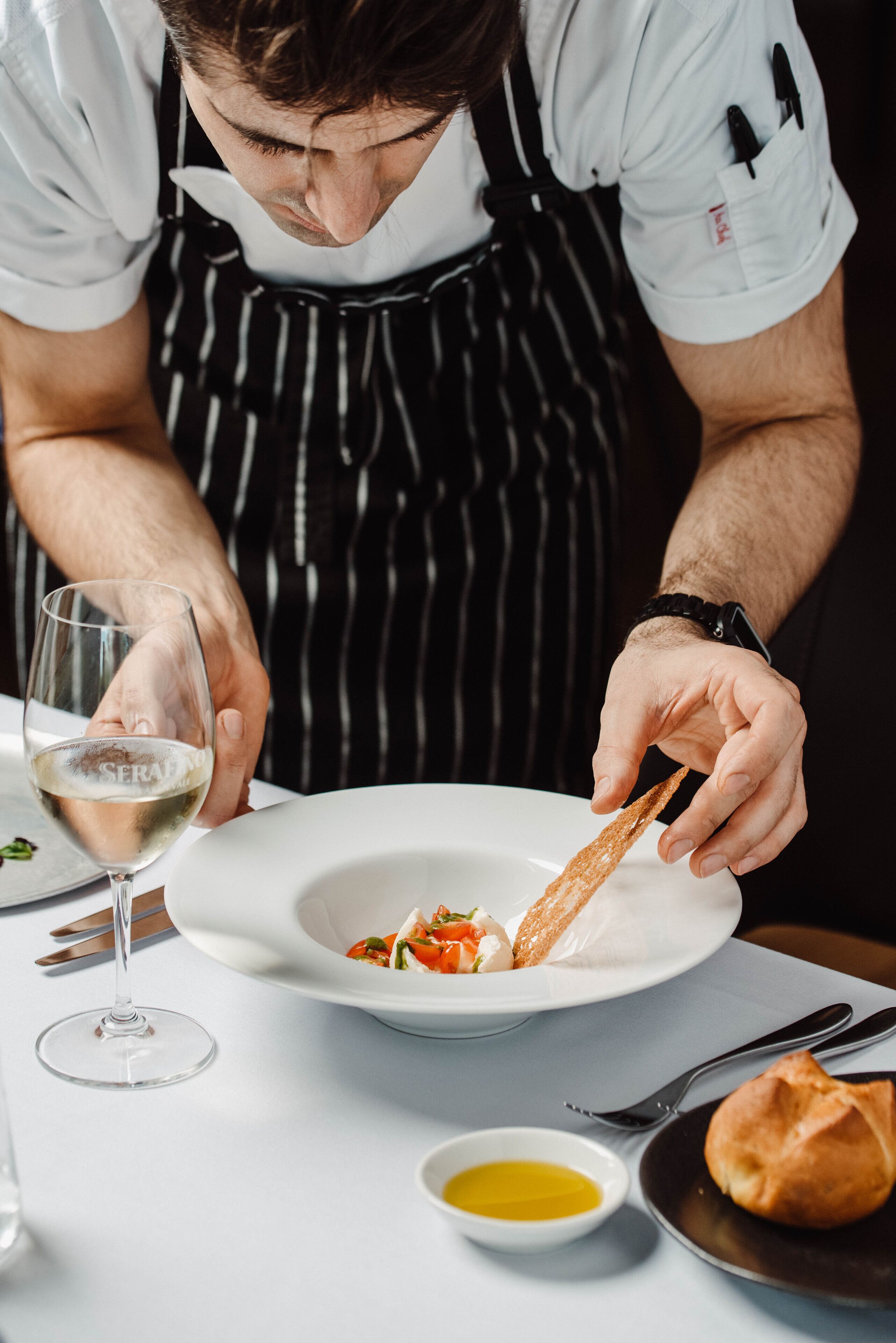 Una persona sirve comida en un plato blanco, con una copa de vino blanco y un recipiente pequeño de aceite de oliva.