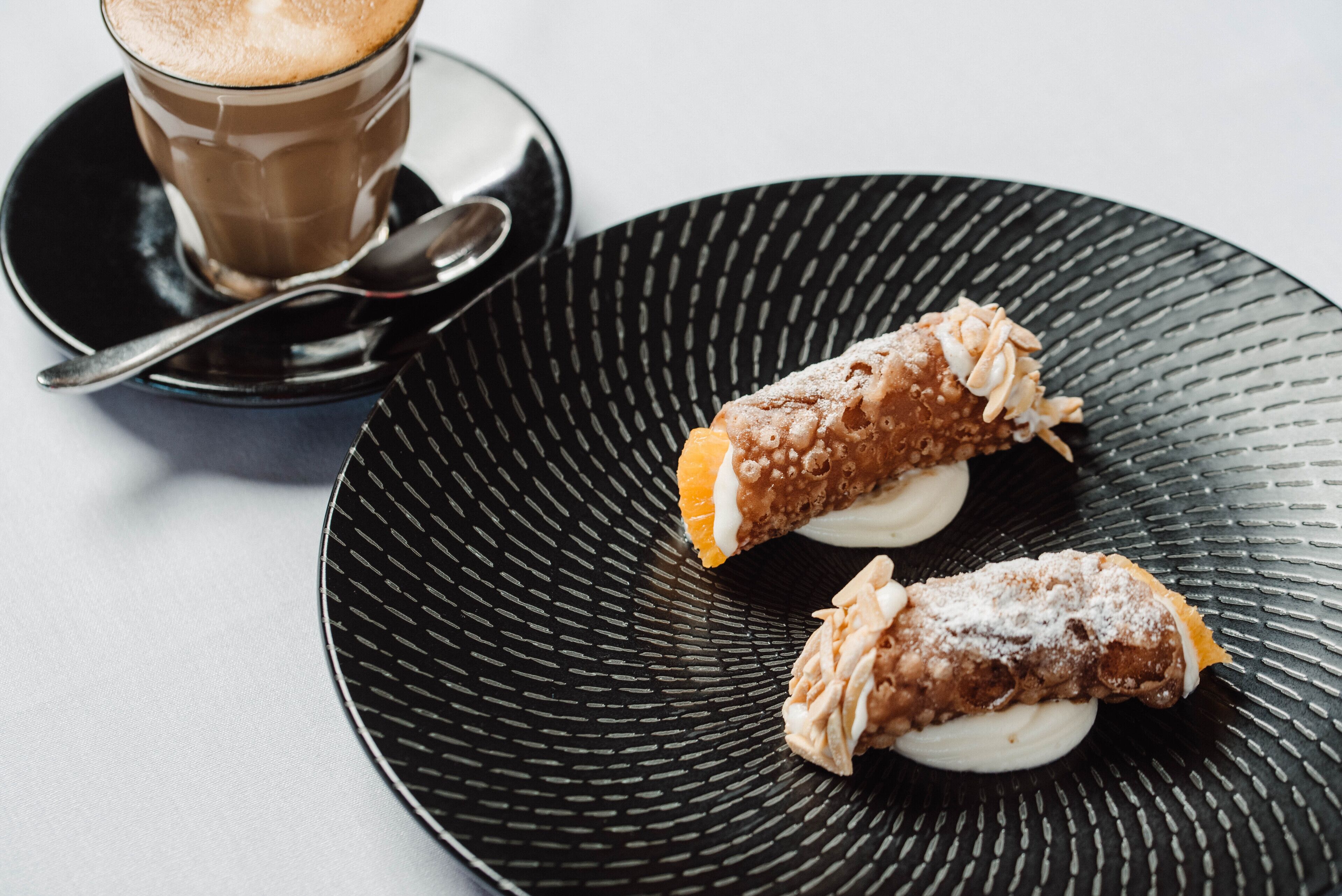 A black plate with two cannoli and a cup of coffee on a saucer.