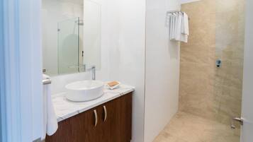 Un baño con un lavamanos blanco, un espejo, una ducha con azulejos color beige y un perchero para toallas.
