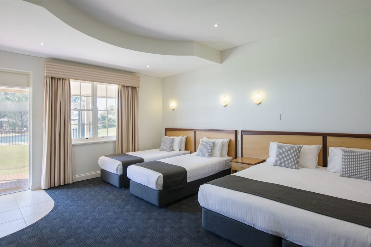 A hotel room with two single beds and one double bed, a large window with curtains, and wall-mounted lights.