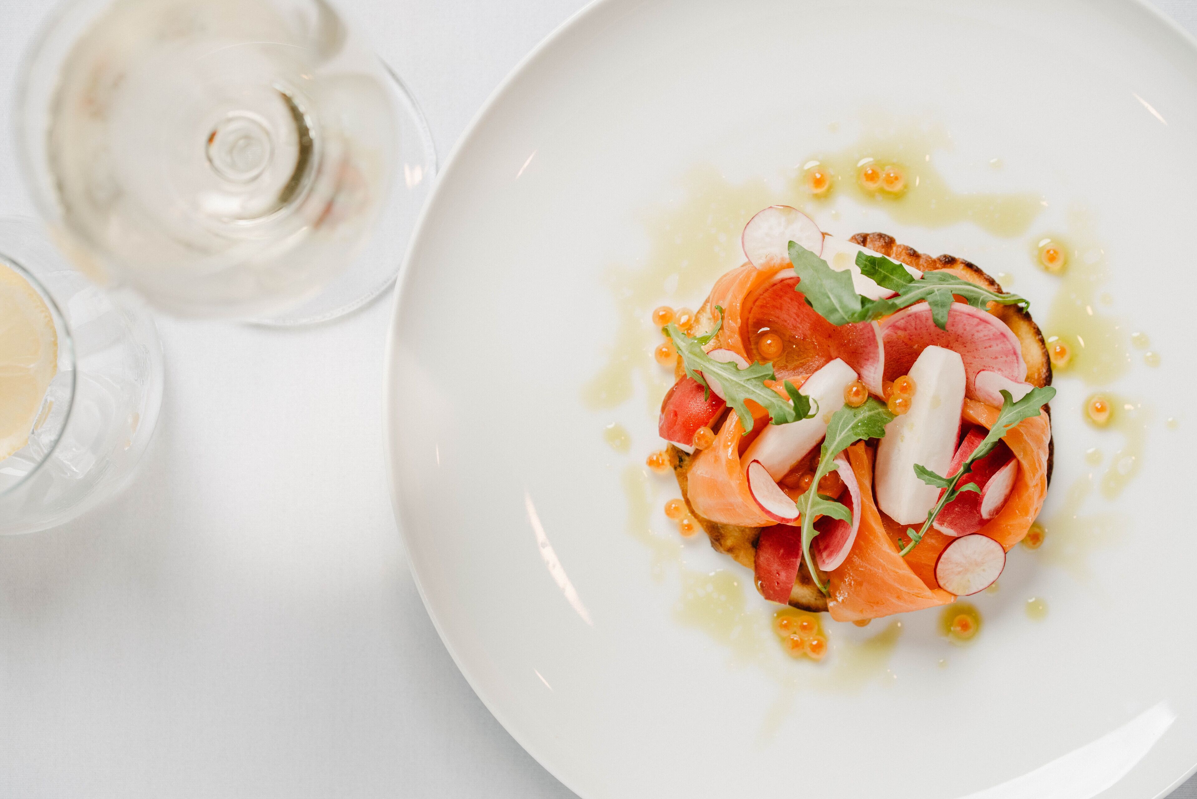 Une assiette blanche contenant des tranches de saumon fumé, des rondelles de radis et des légumes verts, accompagnée d’un verre de vin blanc.