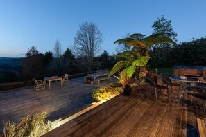 En træterrasse med udendørs møbler, blandt andet borde og stole, omgivet af planter og træer.