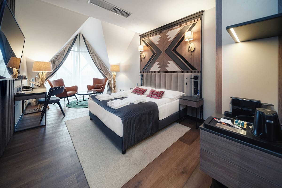 A modern hotel room with a large bed, a desk, and a chair. There is a coffee machine and a TV on the desk.