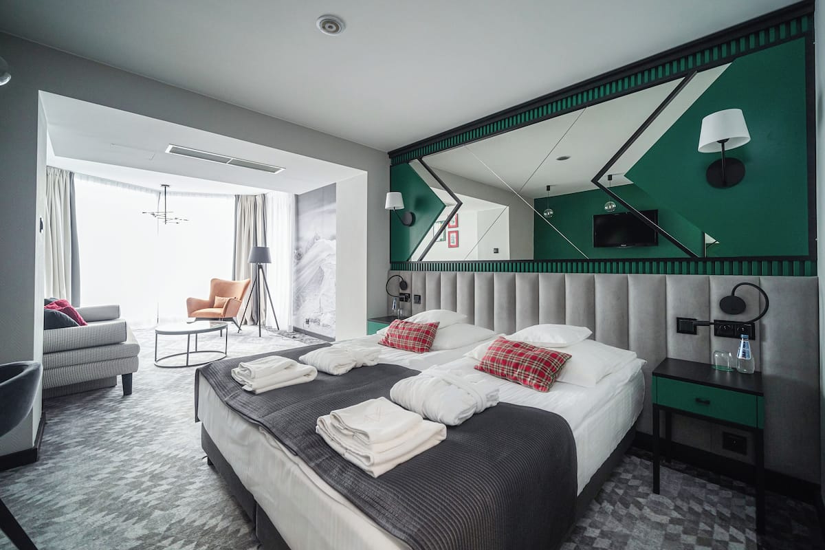 A modern hotel room with a large bed, a seating area with a sofa and armchair, a desk, and a large mirror.
