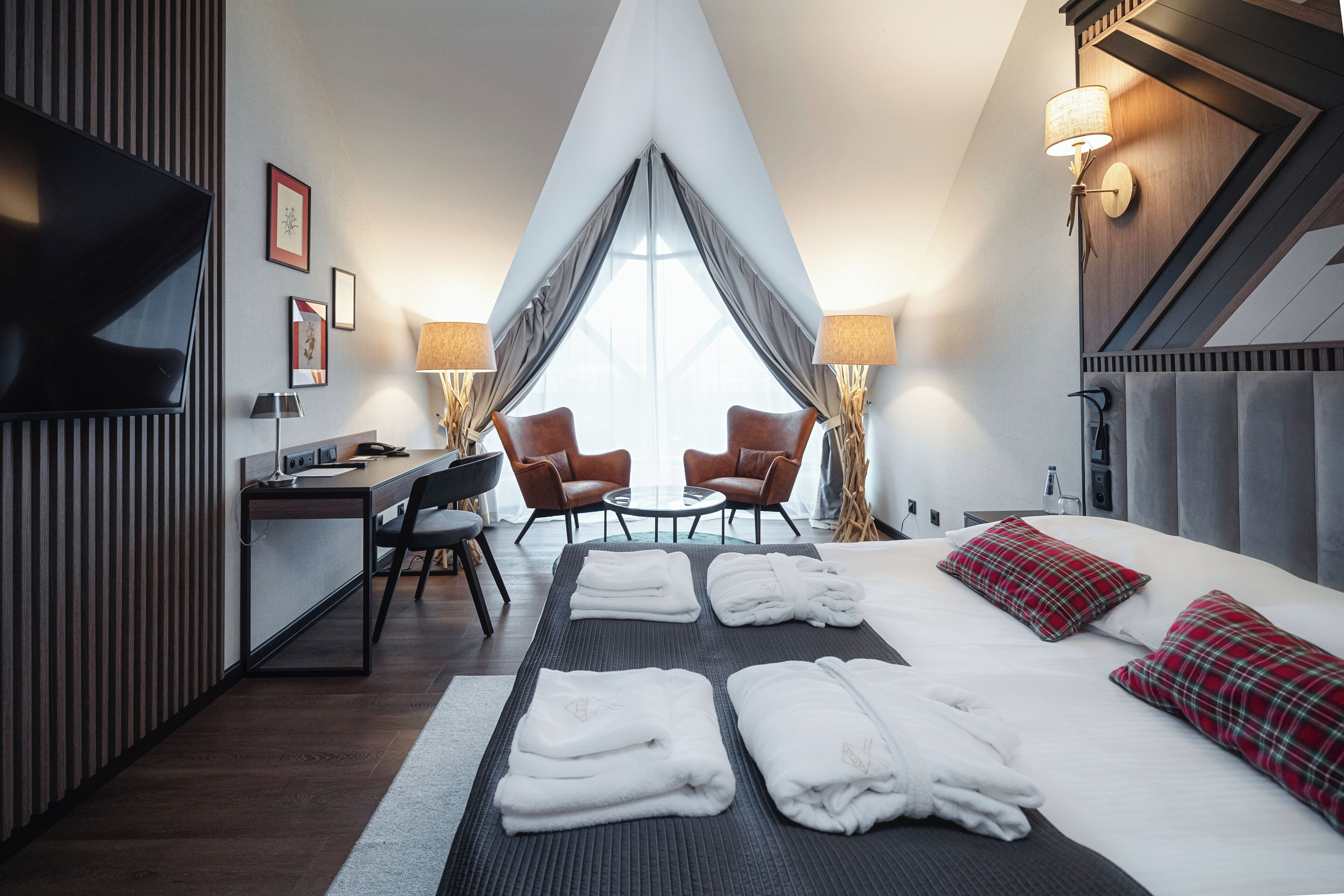 Habitación de hotel con cama, dos sillas, escritorio, televisor y una ventana grande con cortinas.