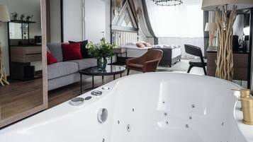 Una habitación de hotel moderna con una bañera blanca grande, un sofá con cojines rojos, una mesa de comedor y un espejo amplio.