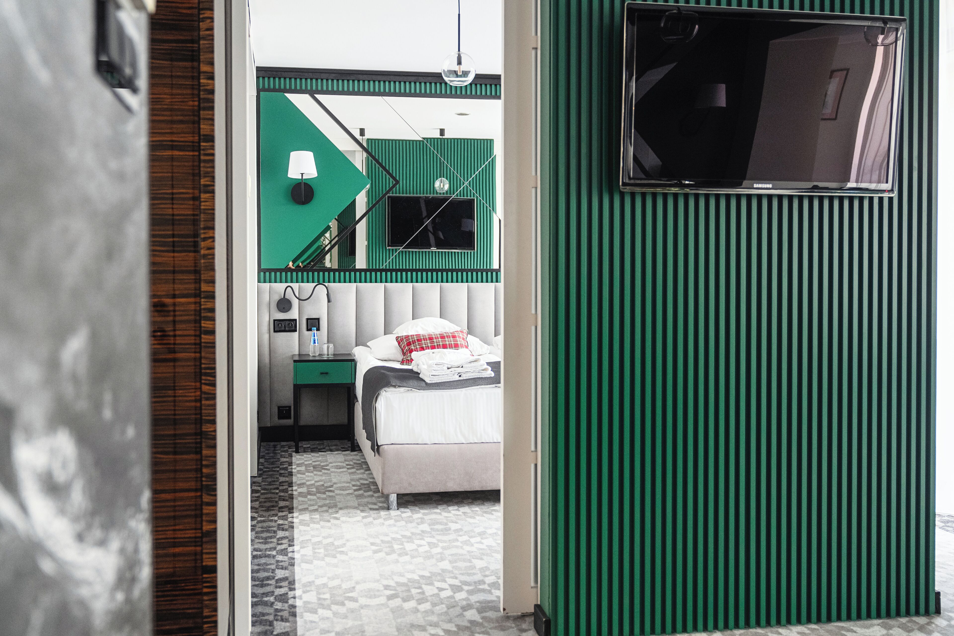 A compact hotel room with a bed, bedside table, lamp, and a green wall with a window.