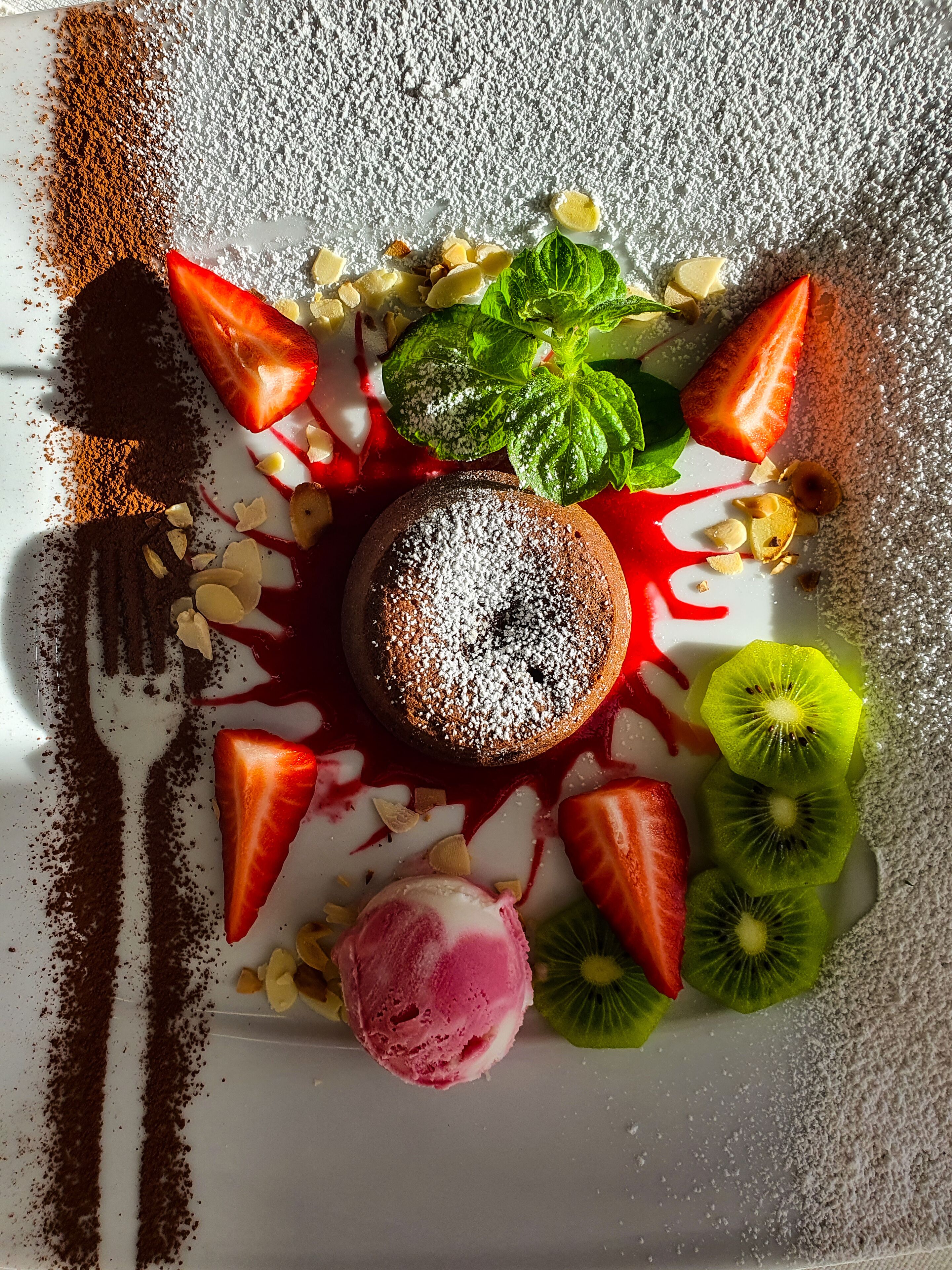 A dessert plate with a chocolate cake, strawberries, kiwi slices, mint leaves, and a scoop of pink ice cream.