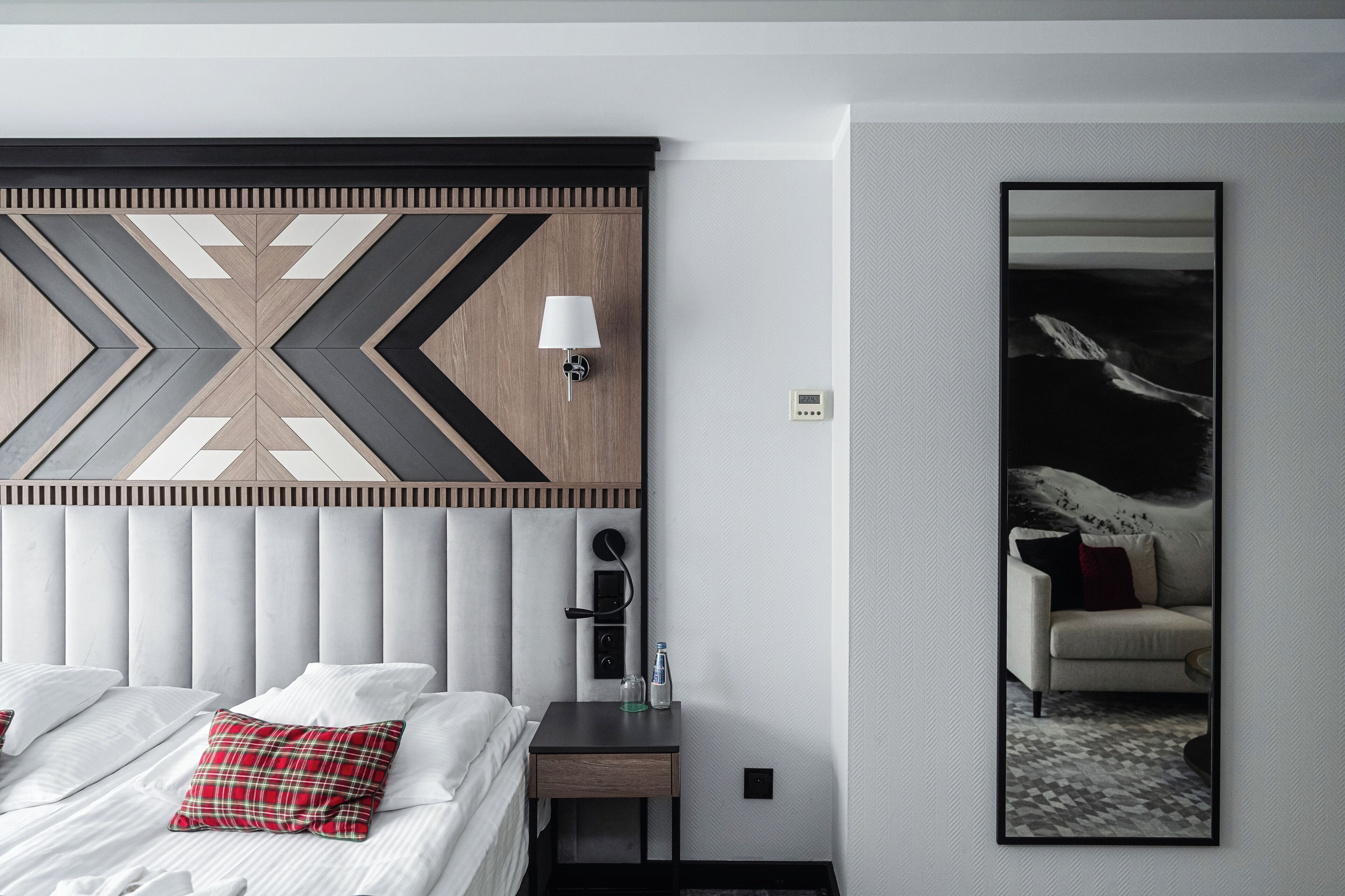 A modern hotel room with a large bed, a bedside table with a lamp, a nightstand, and a large mirror.