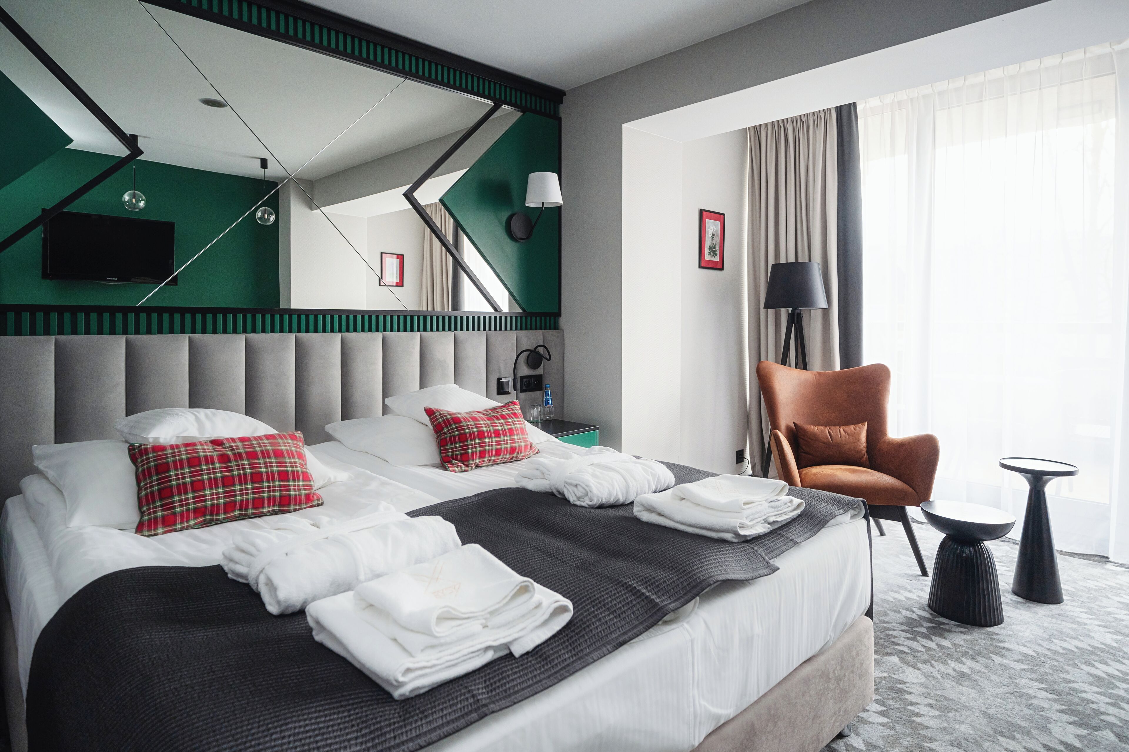 A modern hotel room with a large bed, a grey upholstered headboard, a red plaid pillow, a grey throw blanket, and a brown leather armchair.