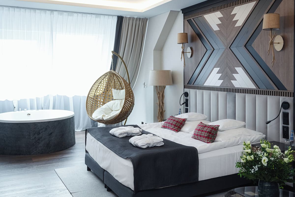 A modern hotel room with a large bed, a round bathtub, a hanging chair, and a decorative wall panel.