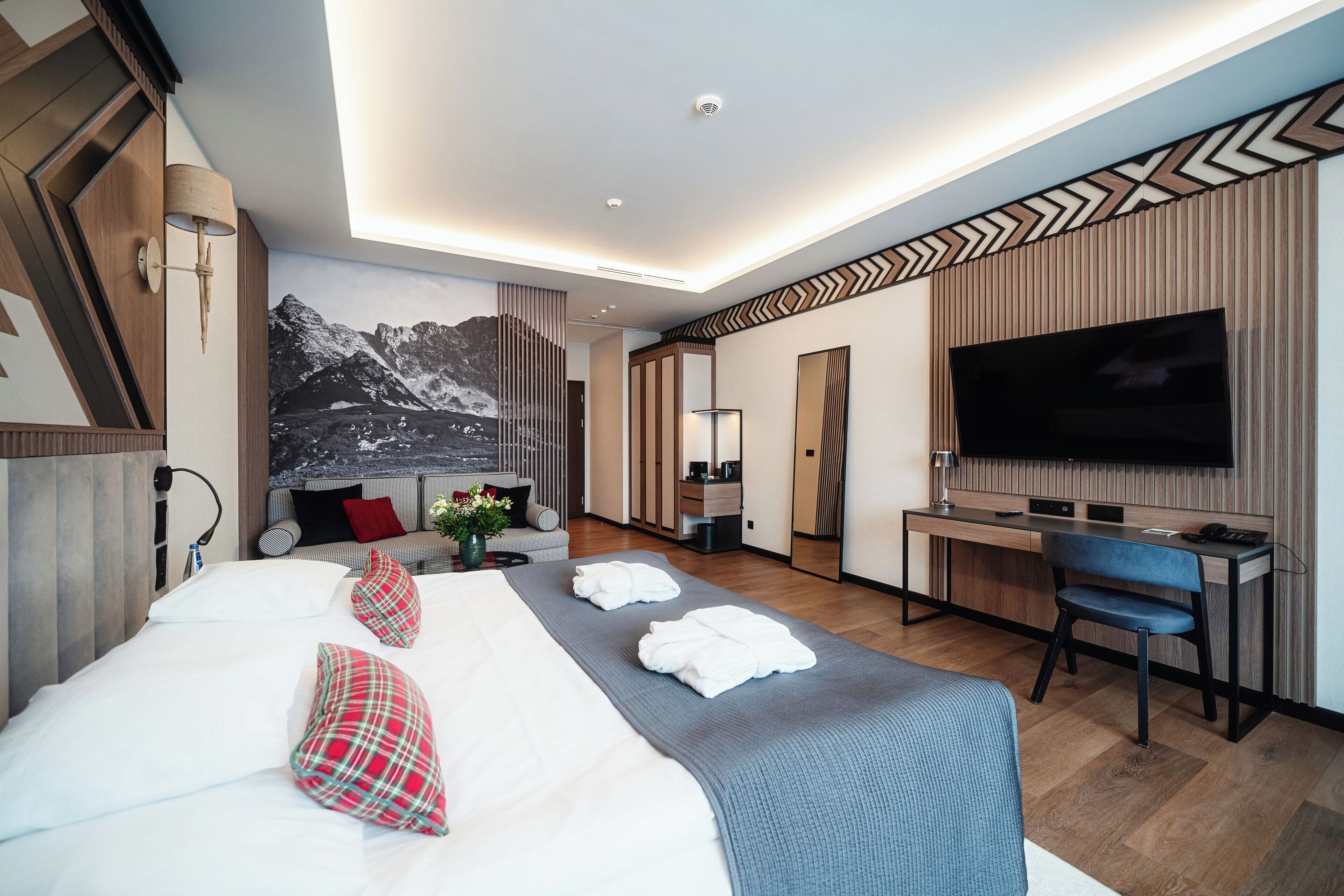 A modern hotel room with a large bed, a desk, a chair, a flat-screen TV, and a mountain mural on the wall.