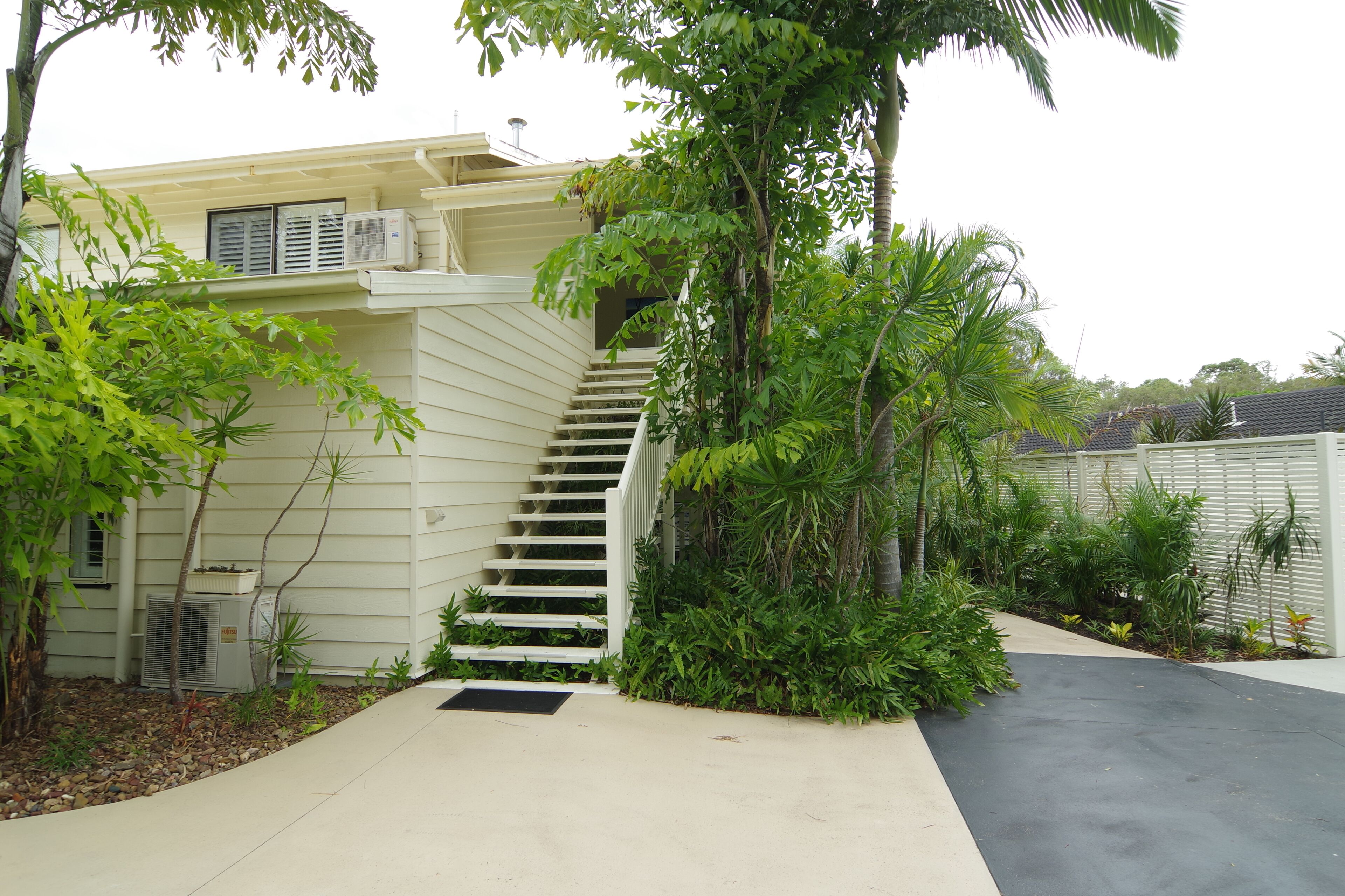 A two-story house with a staircase leading to the upper floor, surrounded by lush greenery and a paved driveway.
