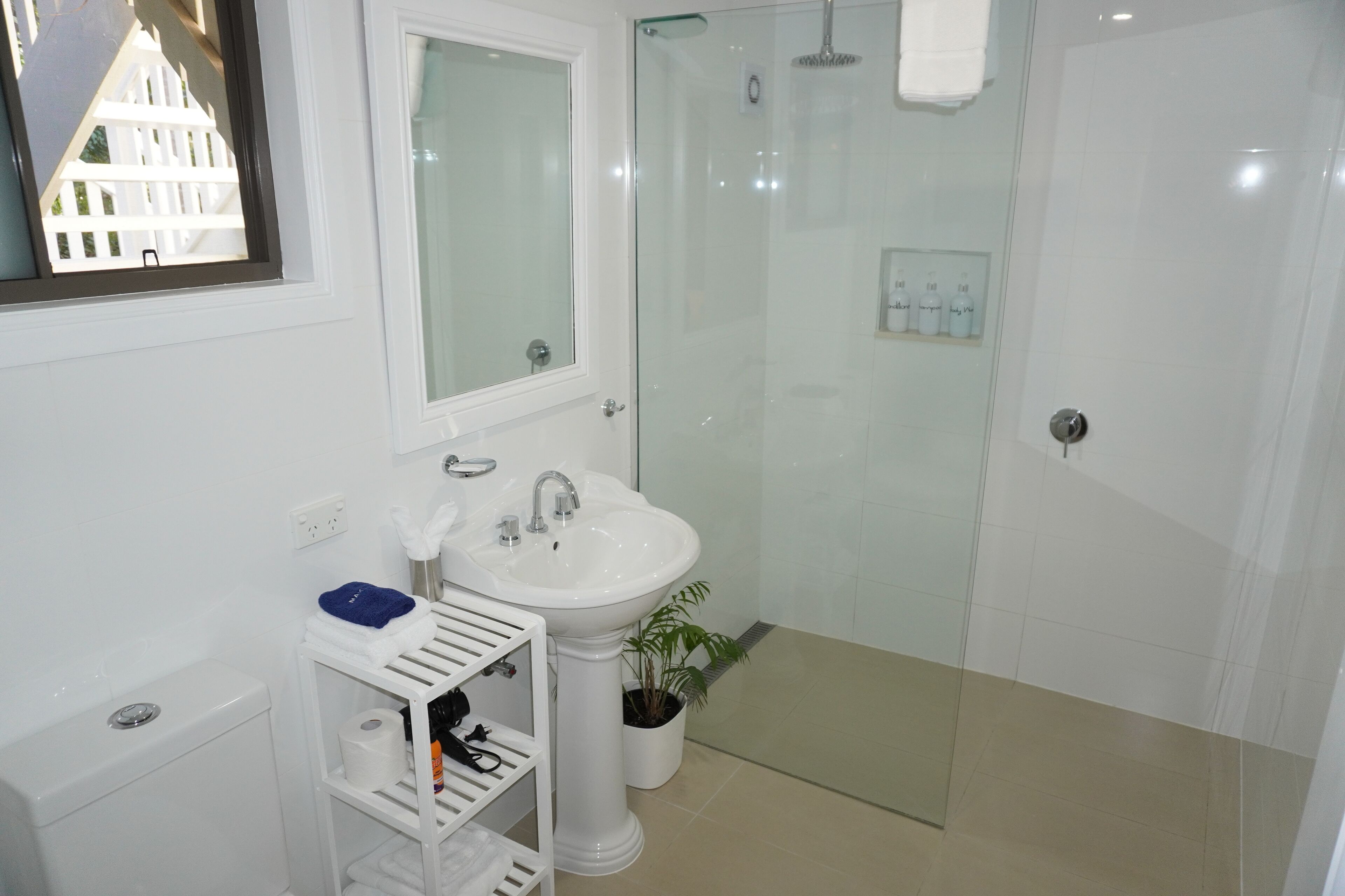 A bathroom with a white sink, mirror, and shower. There is a shelf with toiletries and a potted plant.
