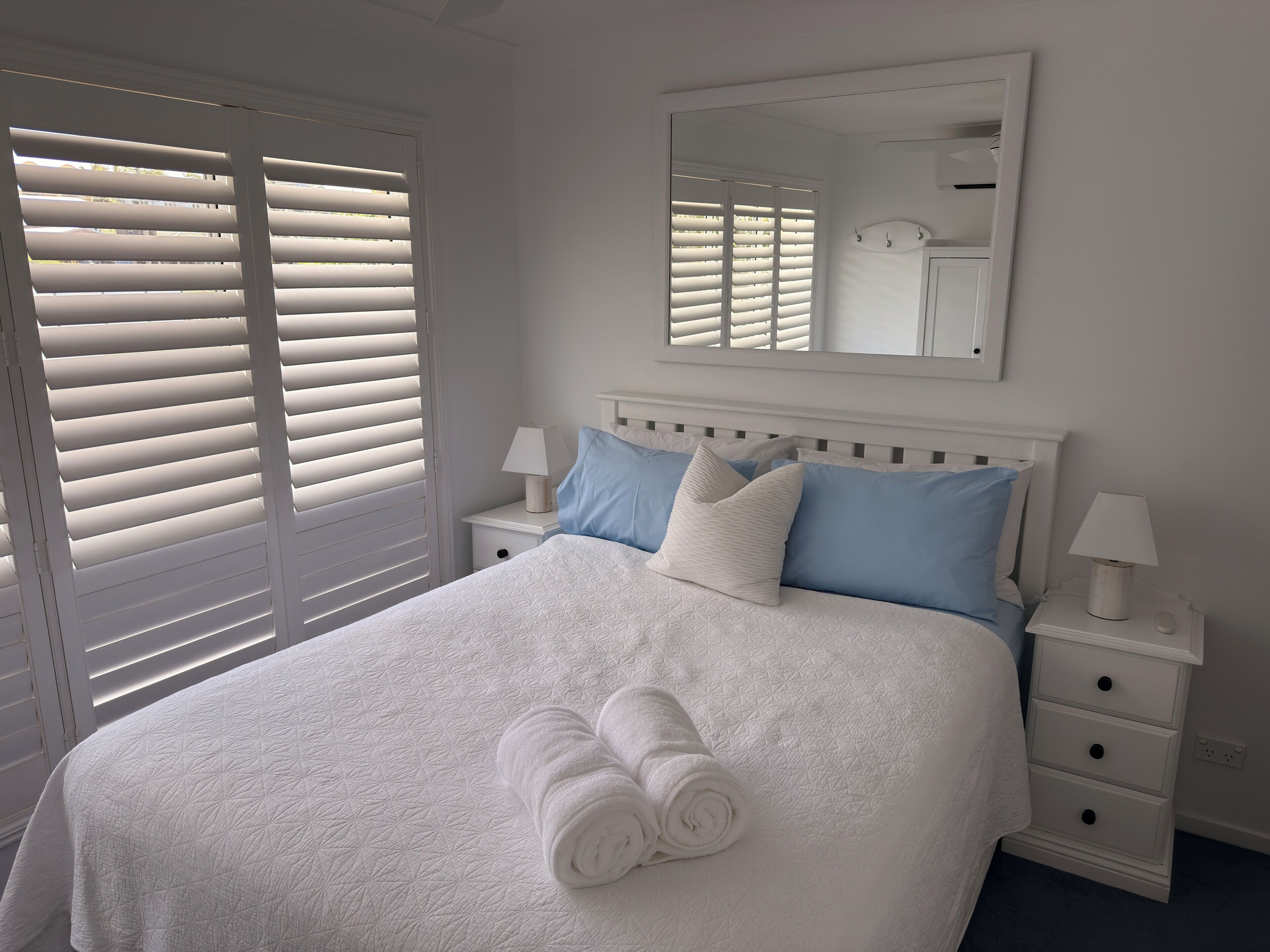 Un letto rifatto con copripiumino bianco trapuntato, due cuscini blu e bianchi, e due asciugamani bianchi arrotolati. Si nota un comodino bianco con una lampada e uno specchio appeso alla parete sopra il letto.