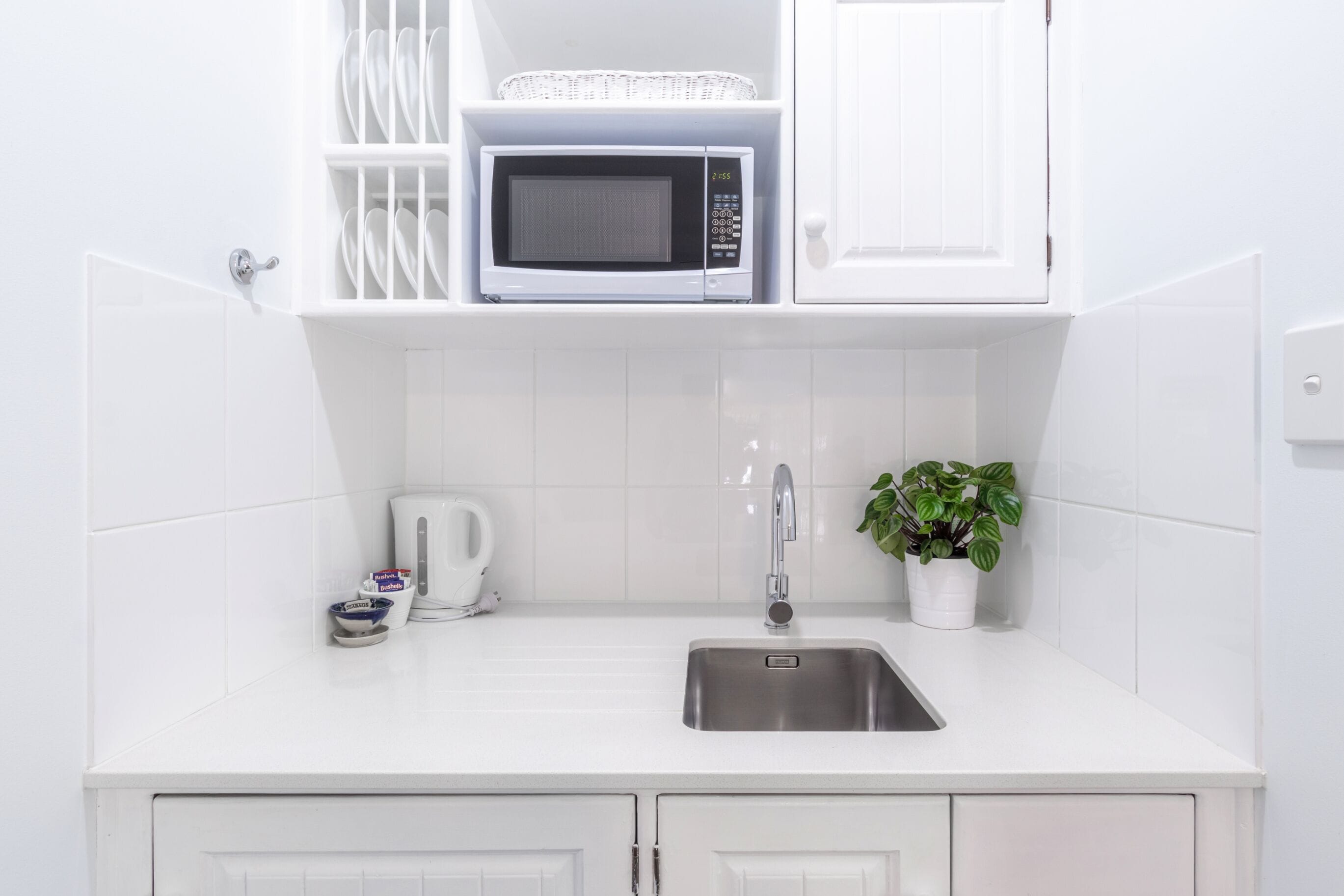 Eine moderne Küche mit weißen Schränken, einer Spüle aus Edelstahl, einer Mikrowelle, einem Wasserkocher und einem Blumentopf.