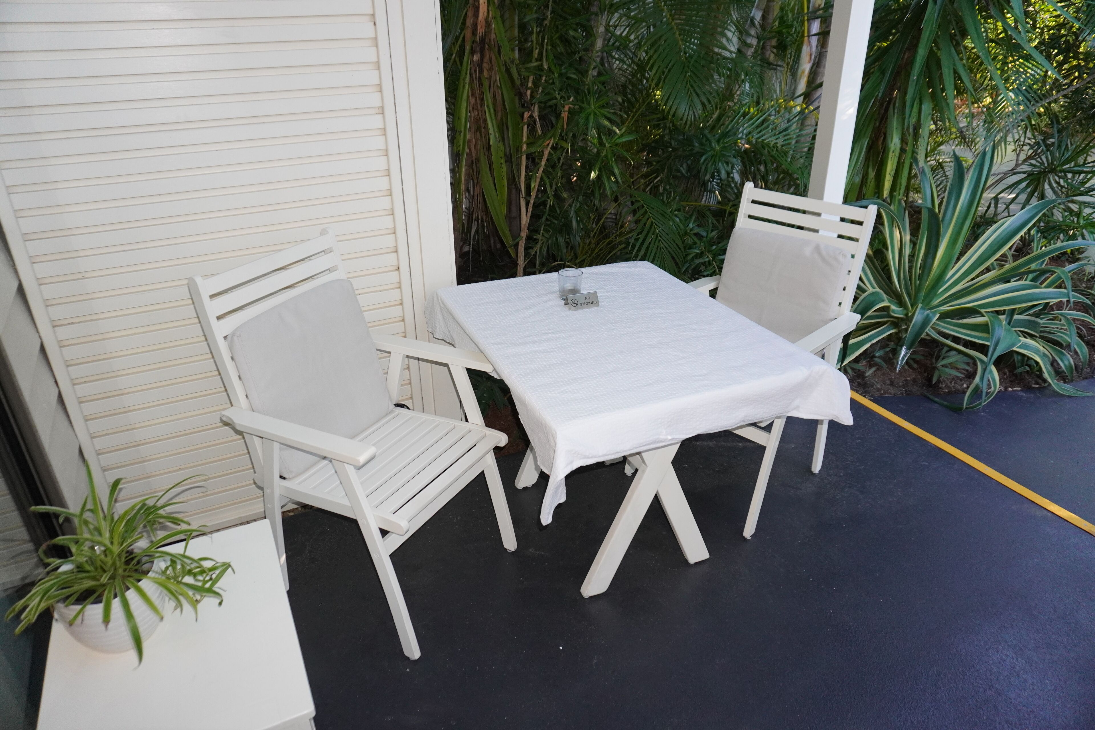 Une table blanche et deux chaises blanches sont disposées sur un balcon offrant une vue sur une végétation luxuriante.