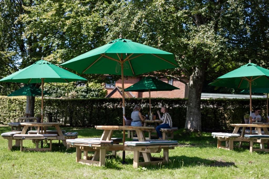 Outdoor wooden picnic tables with green umbrellas, surrounded by trees and a hedge.