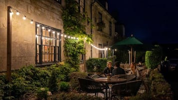 An outdoor seating area with tables and chairs, surrounded by greenery and a building facade.